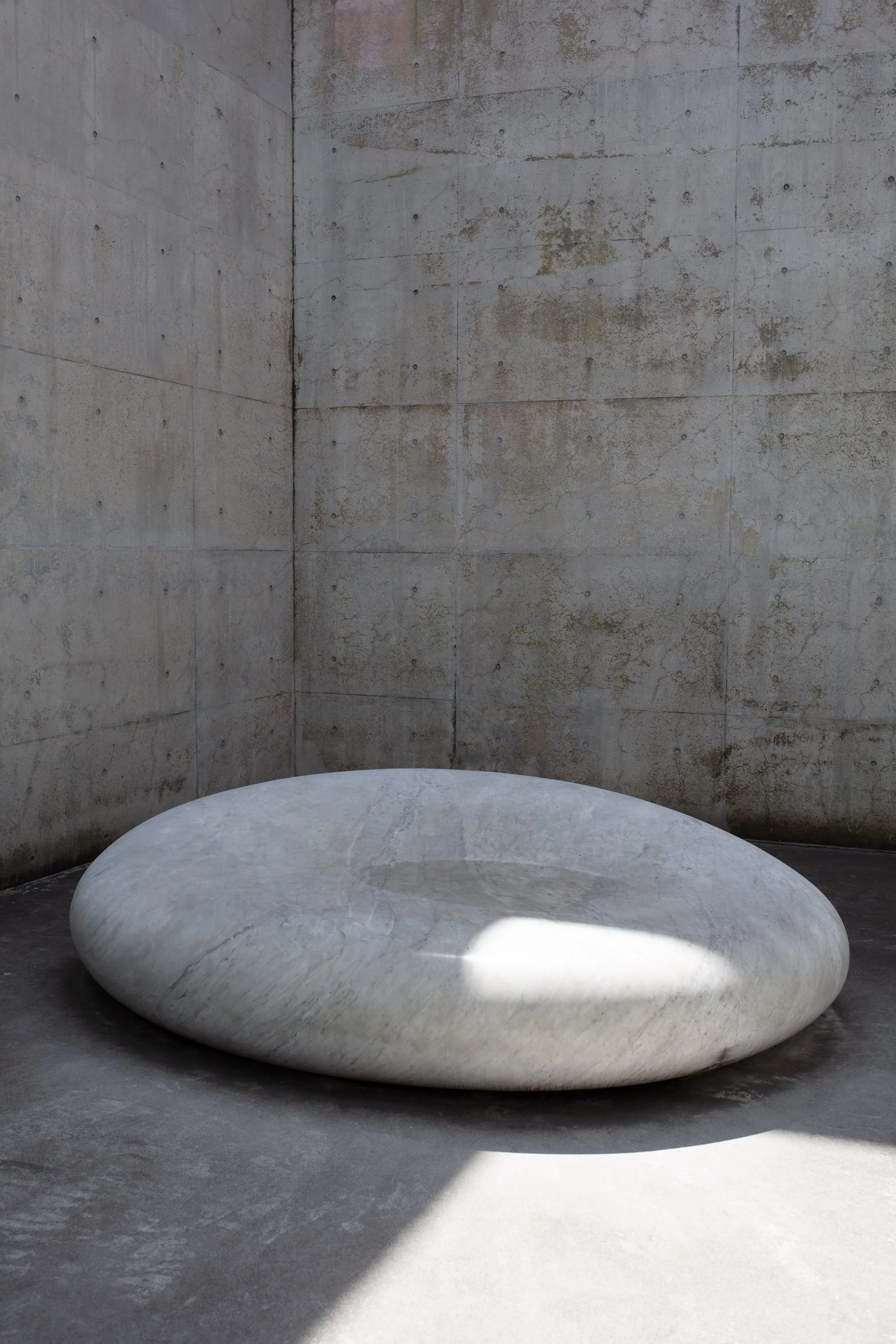 A large, smooth, oval-shaped white marble stone in a corner with gray concrete walls and floor. Sunlight casts a shadow and a bright reflection on the stone.