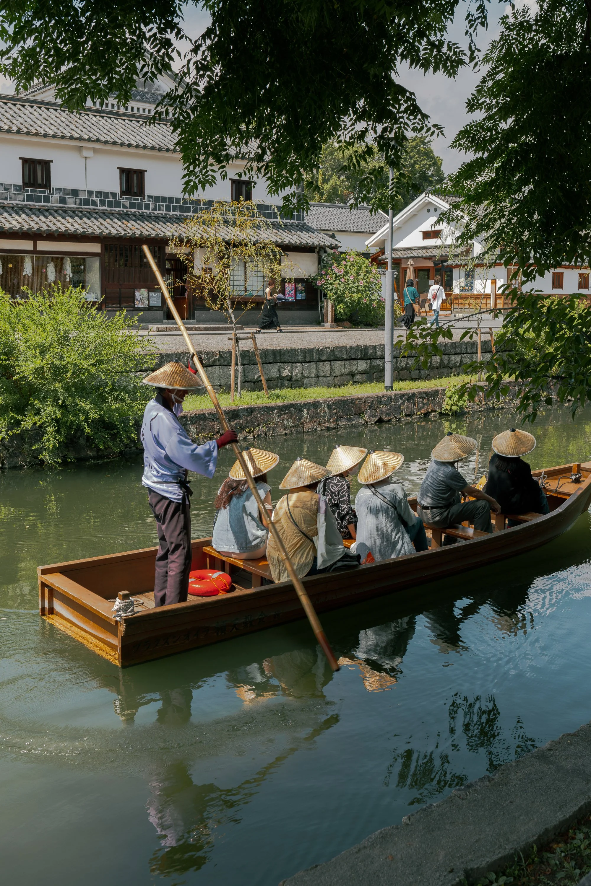 People in traditional attire and conical hats taking a boat ride along a river surrounded by greenery, with traditional buildings in the background.