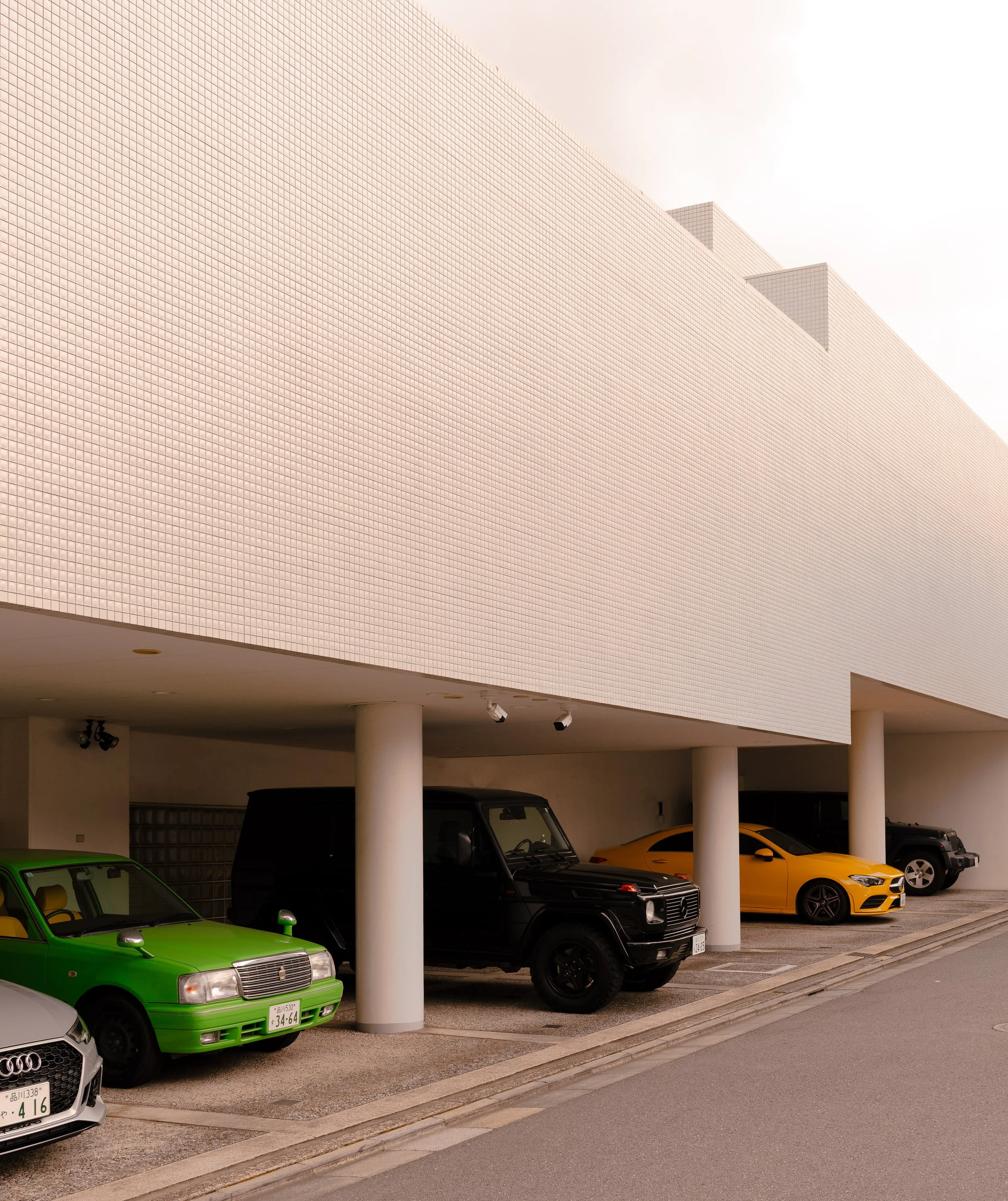 Undercover parking garage with a variety of parked cars, including a green car, a black SUV, a yellow sports car, and a gray vehicle, beneath a large modern building with white tiled exterior and round columns supporting the structure.