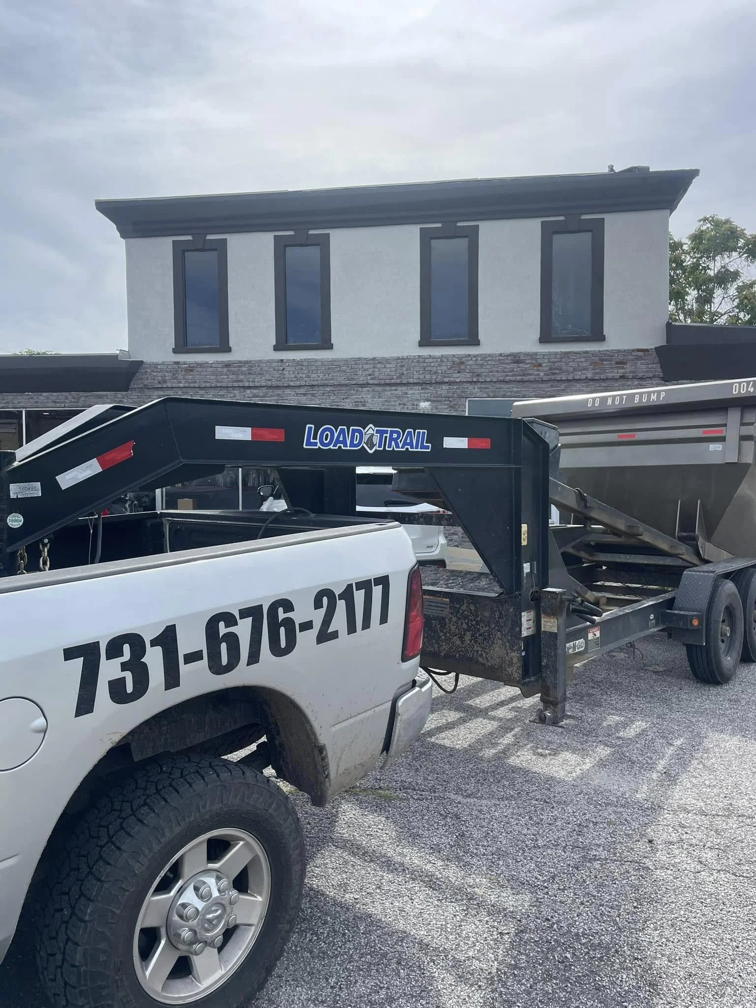 A pickup truck with a large load trail attached, parked in front of a modern white building with four tall windows and a brick section below the windows, under a cloudy sky.