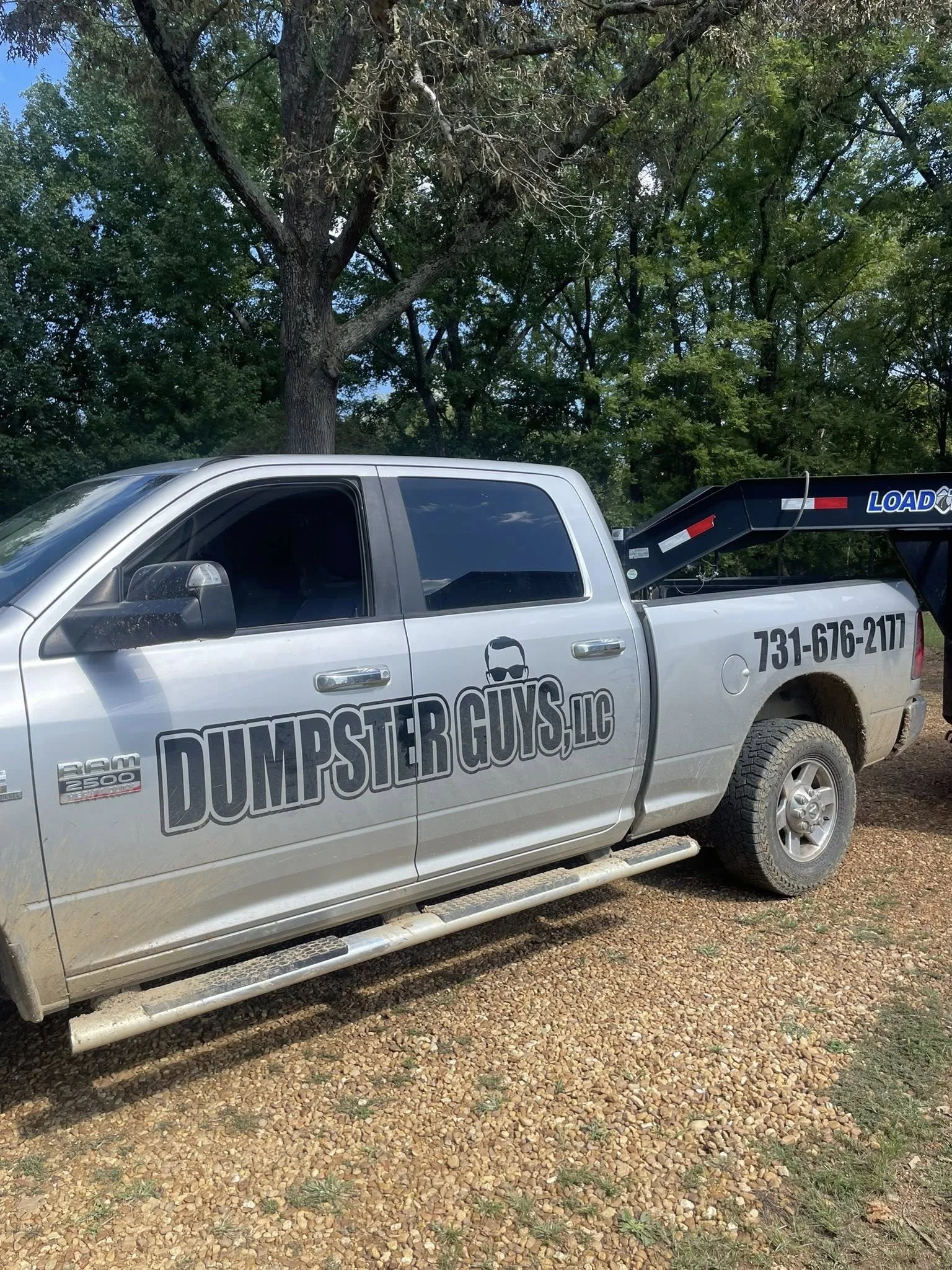A silver pickup truck with the logo and name "Dumpster Guys LLC" on the side, parked on a gravel surface in front of trees.