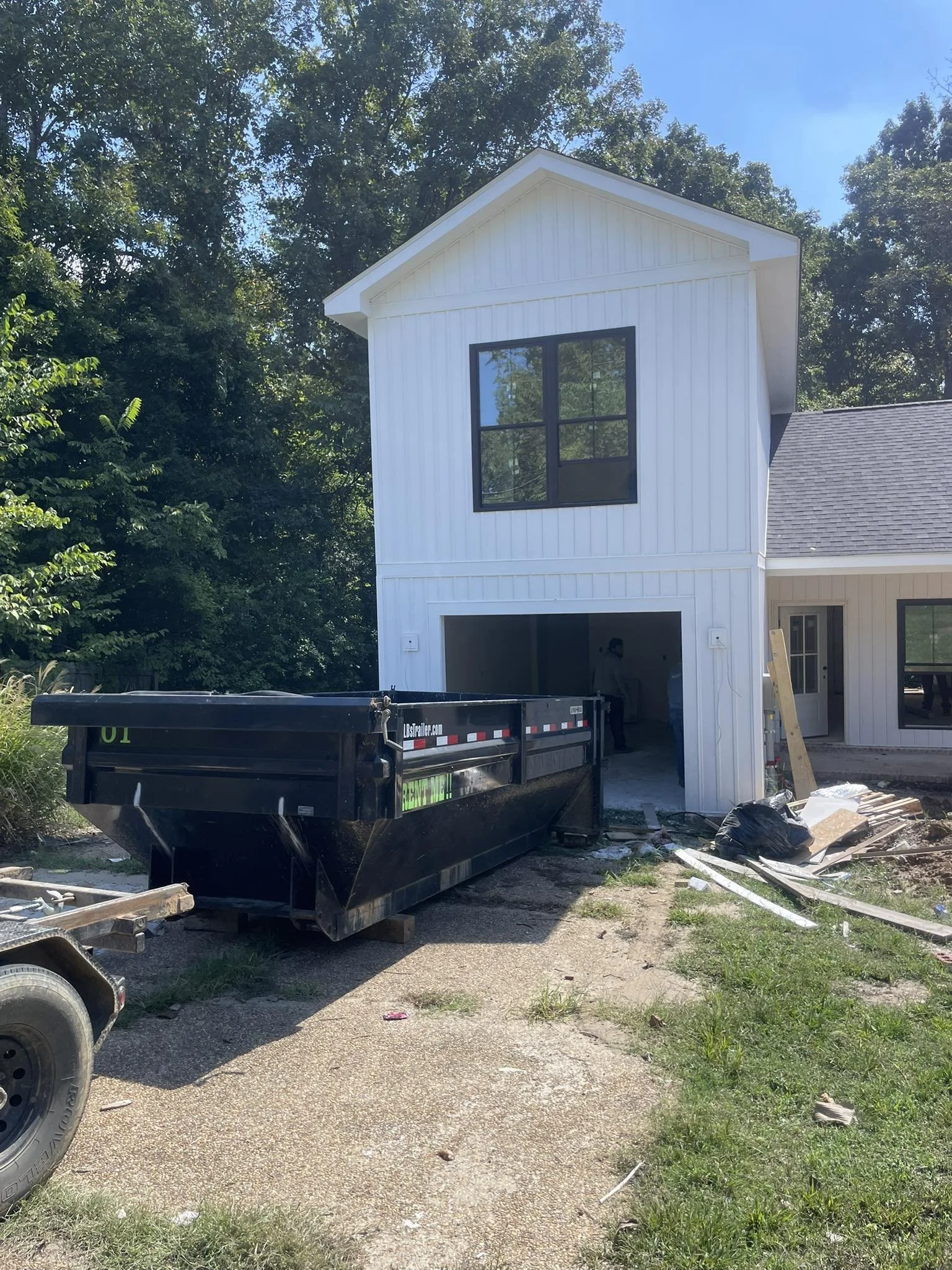 Under construction house with white siding, large window, and an open garage. A black dump trailer is parked in front, and construction materials are scattered around.