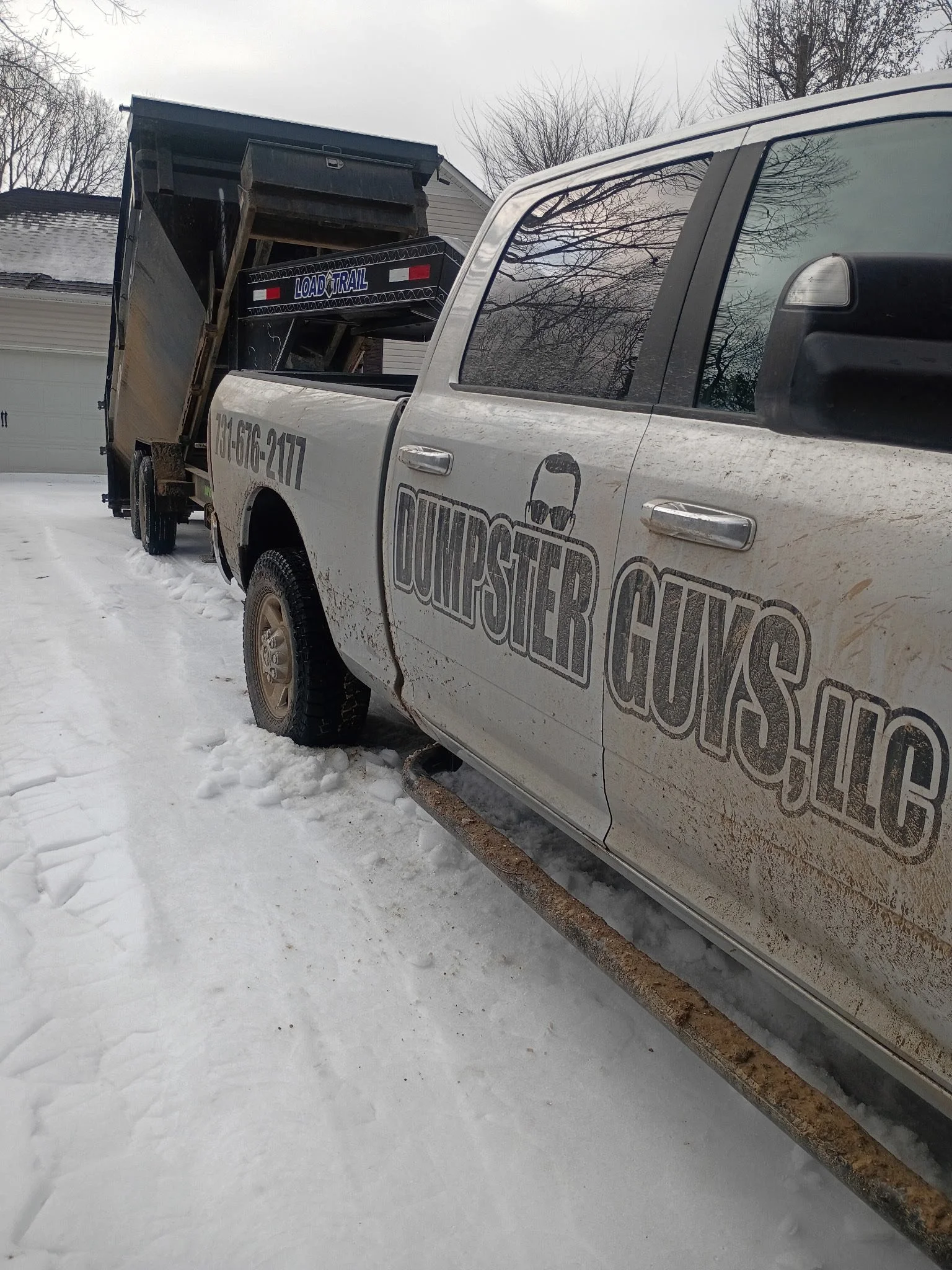 A white pickup truck with a logo and text that says 'DUTPSTER GUYS' on the door, parked on a snowy driveway. The truck is carrying a trailer with a large black item inside, and the background shows some houses and leafless trees.