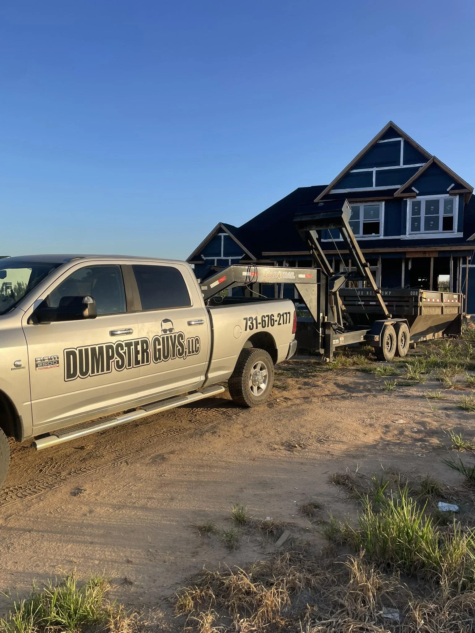 A pickup truck labeled 'Dumpster Guys LLC' is towing a construction trailer in front of a house under construction in a rural area with grass and dirt ground.