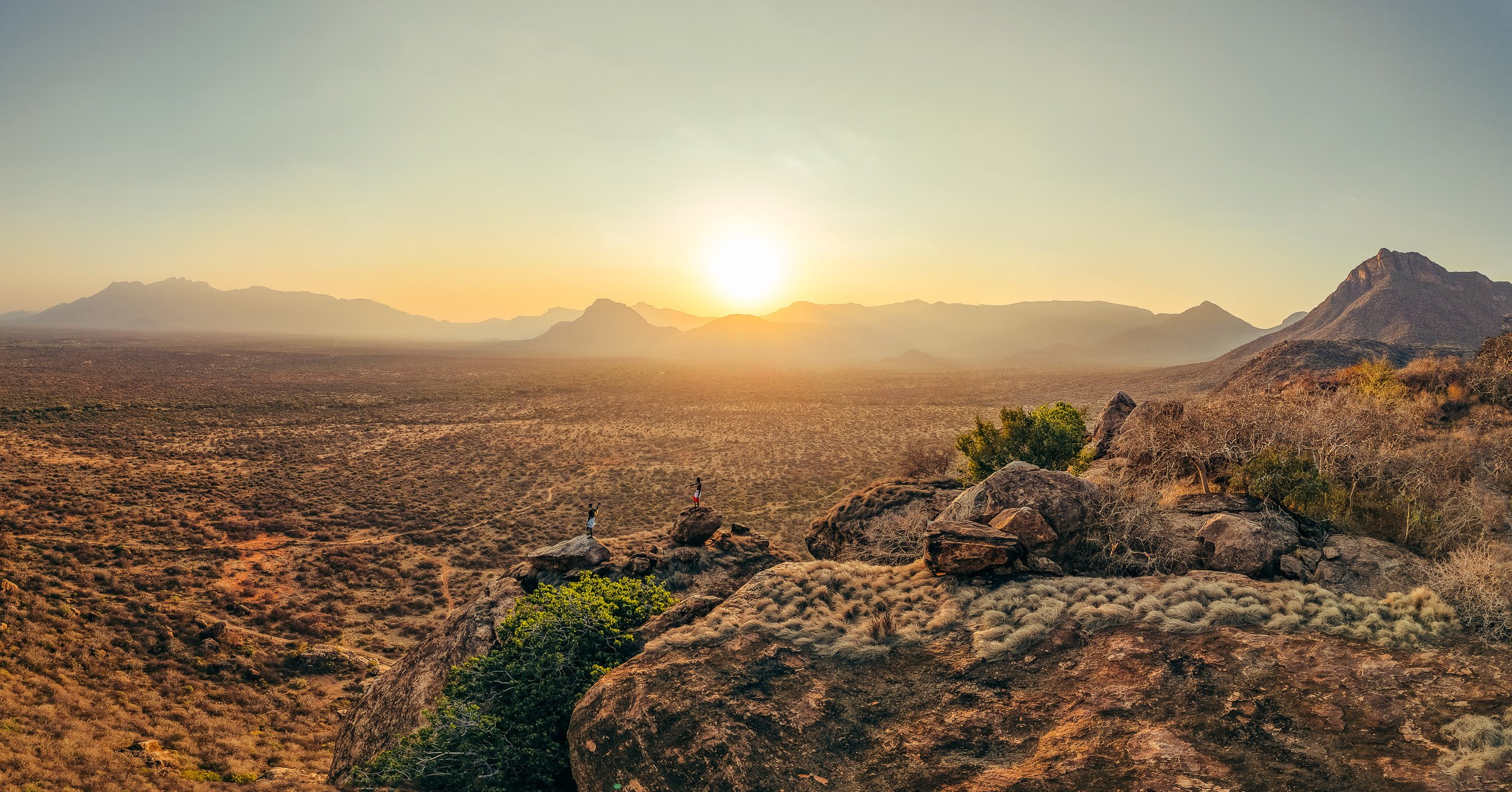 Desert landscape at sunset with mountains in the distance, large rocks and sparse vegetation in the foreground, two people walking on rocks.
