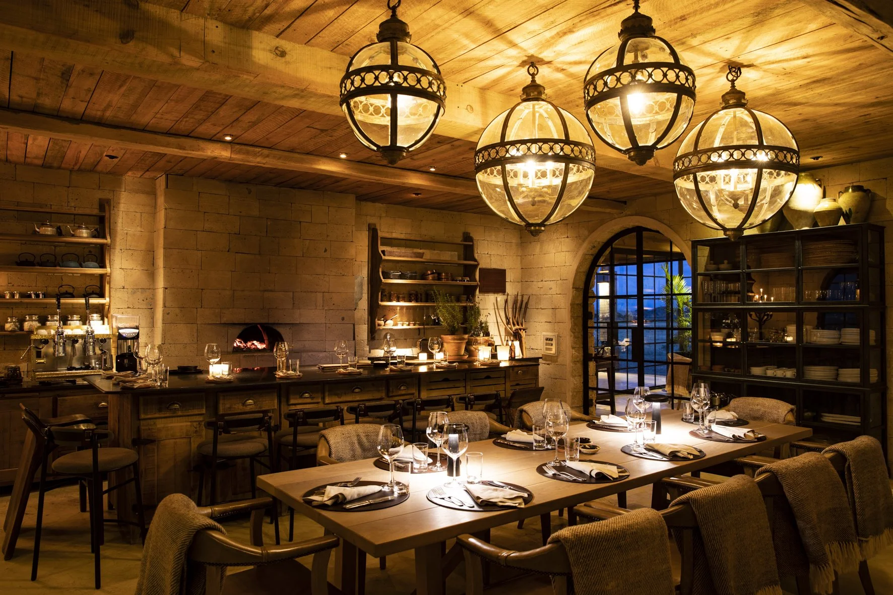 A cozy, warmly lit dining area with rustic decor. The room features a long wooden table set with glasses, napkins, and plates, surrounded by upholstered chairs. Overhead, four large, round, vintage-style pendant lights hang from the wooden ceiling. The back wall has open shelving with dishes and decorative items, a small fireplace, and a large arched window that reveals a patio with outdoor seating and greenery outside.