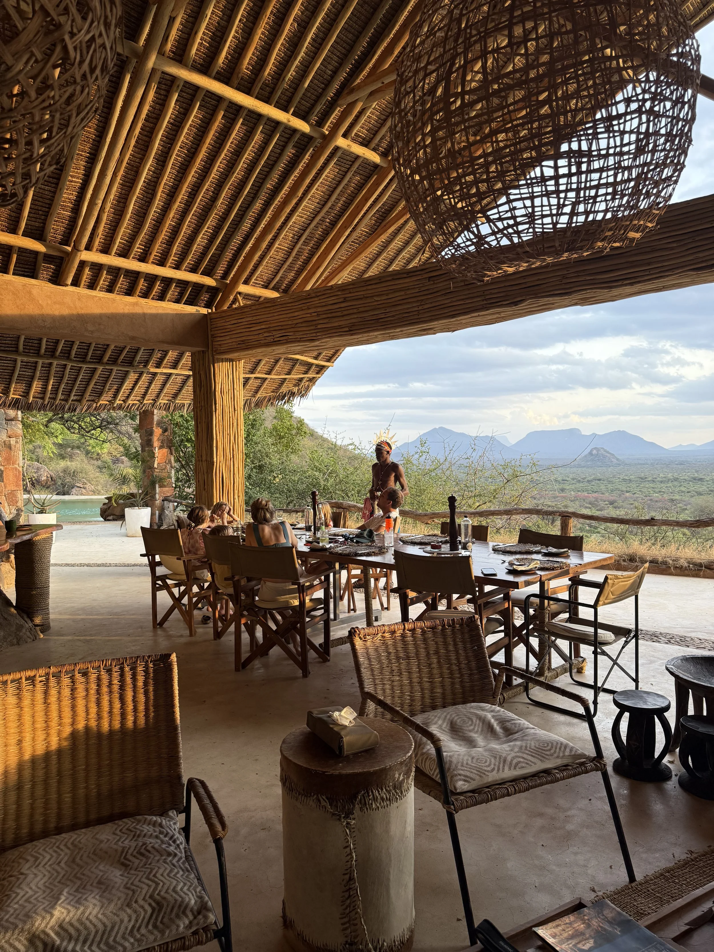 An outdoor dining area with wooden furniture and a thatched roof, overlooking a scenic landscape with mountains and greenery in the background. There are several people seated at the table, and a person standing nearby, possibly during a meal or gathering.