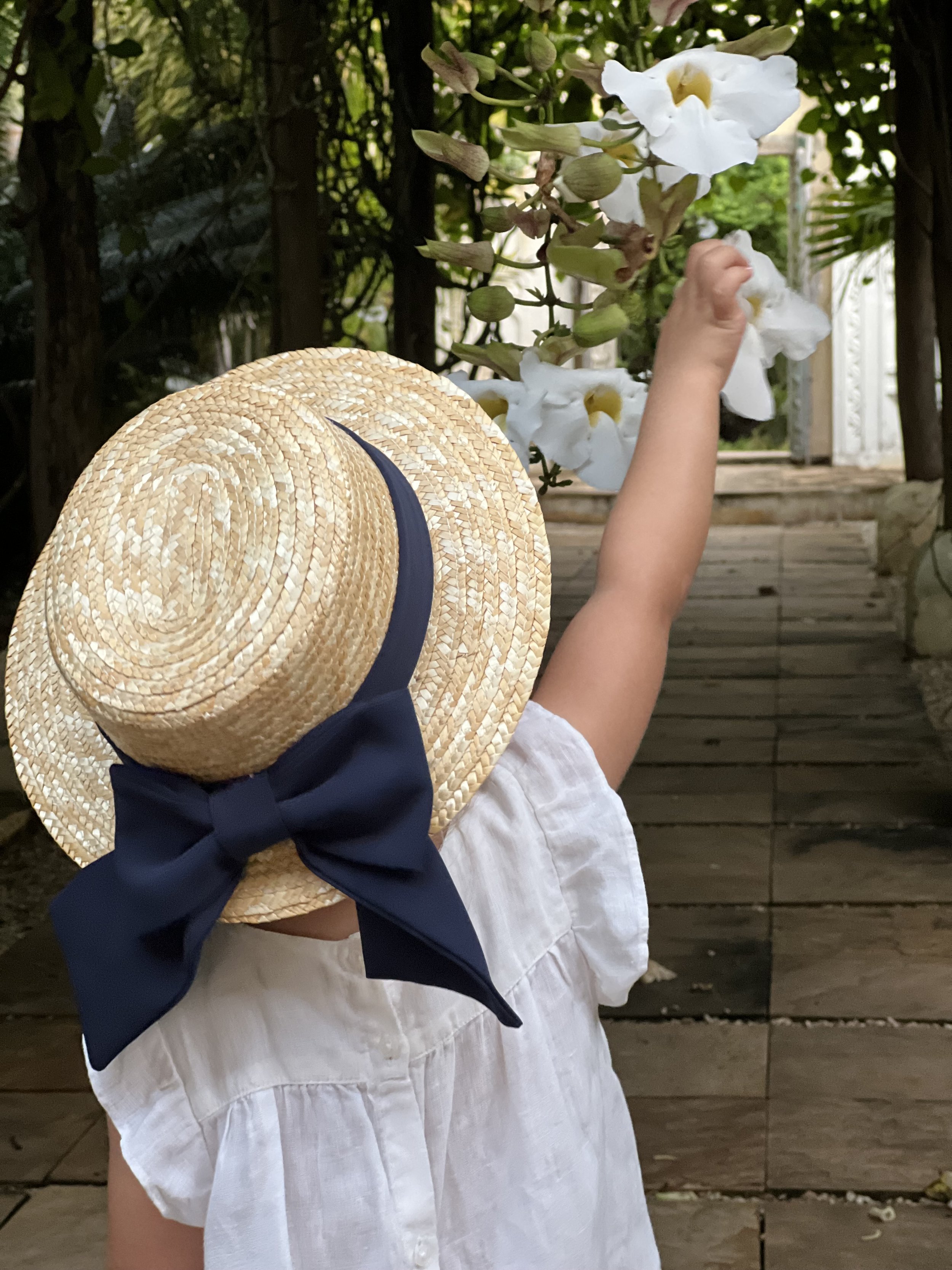 Child reaching up to touch white flowers with a straw sun hat decorated with a navy blue bow, standing on a wooden pathway in a garden.