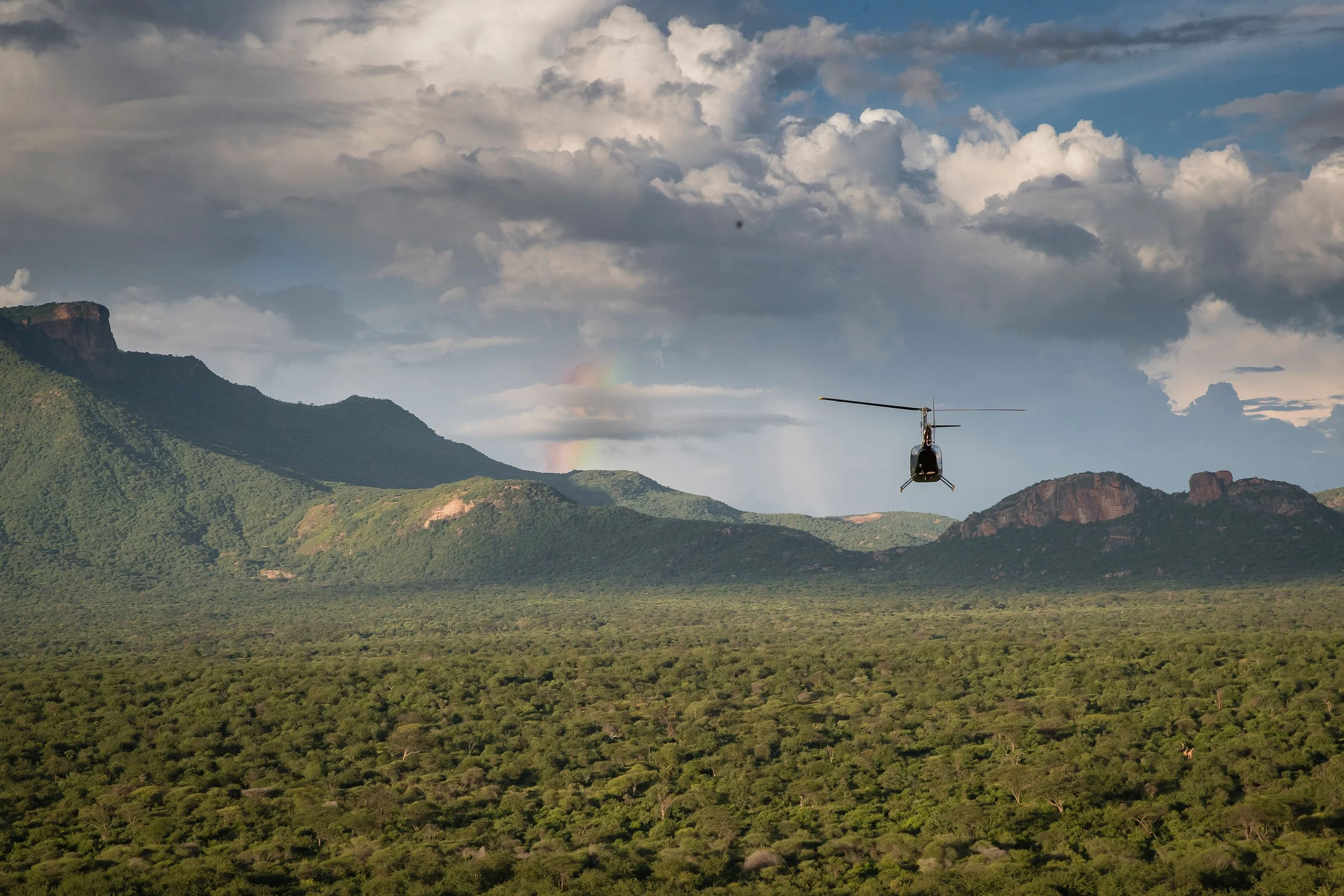 A helicopter flying over a lush, green landscape with mountains in the background and a sky filled with clouds and a faint rainbow.