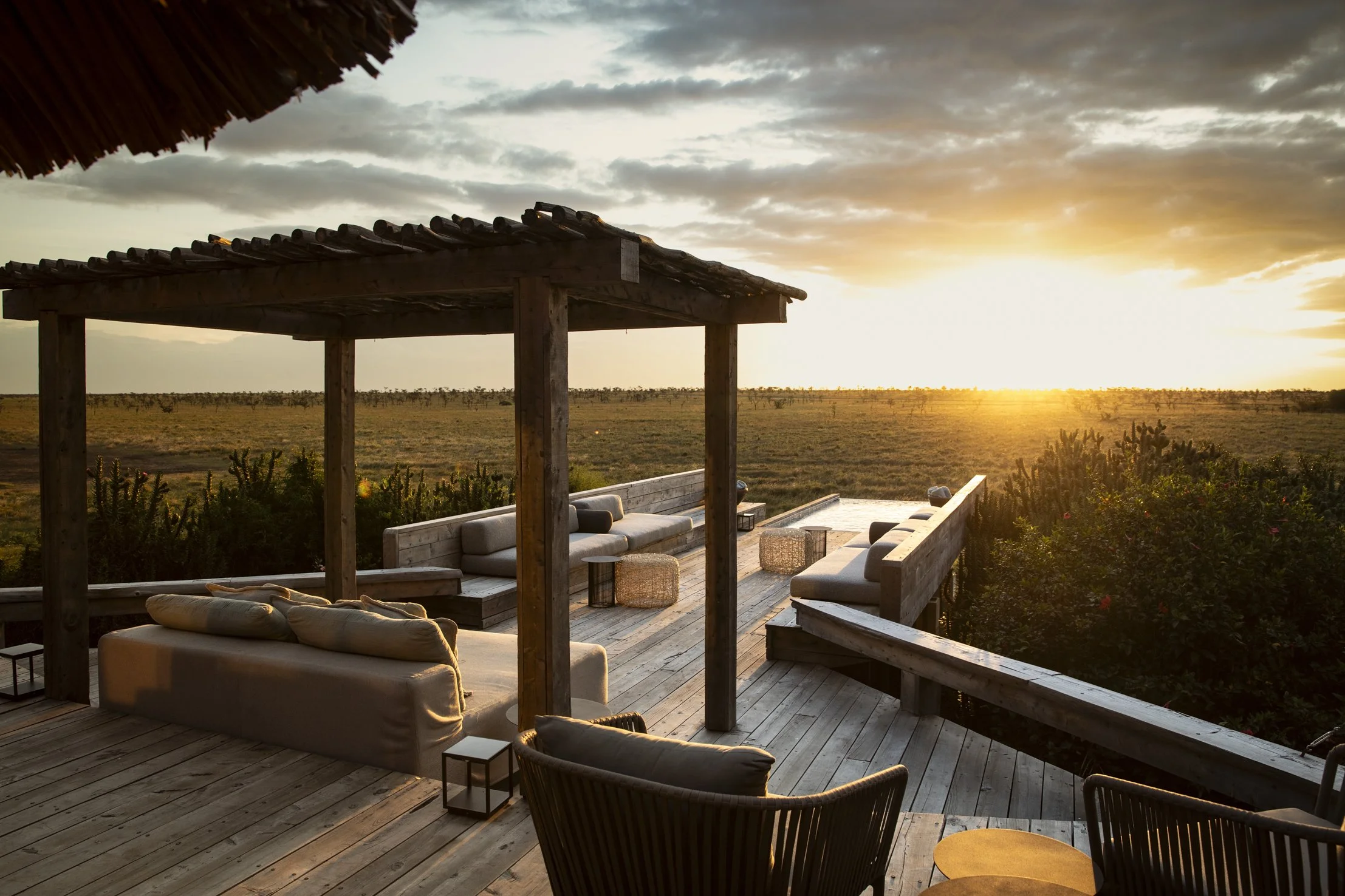 Sunset view from a wooden deck with outdoor furniture including sofas, chairs, and small tables, overlooking a vast open field and cloudy sky.