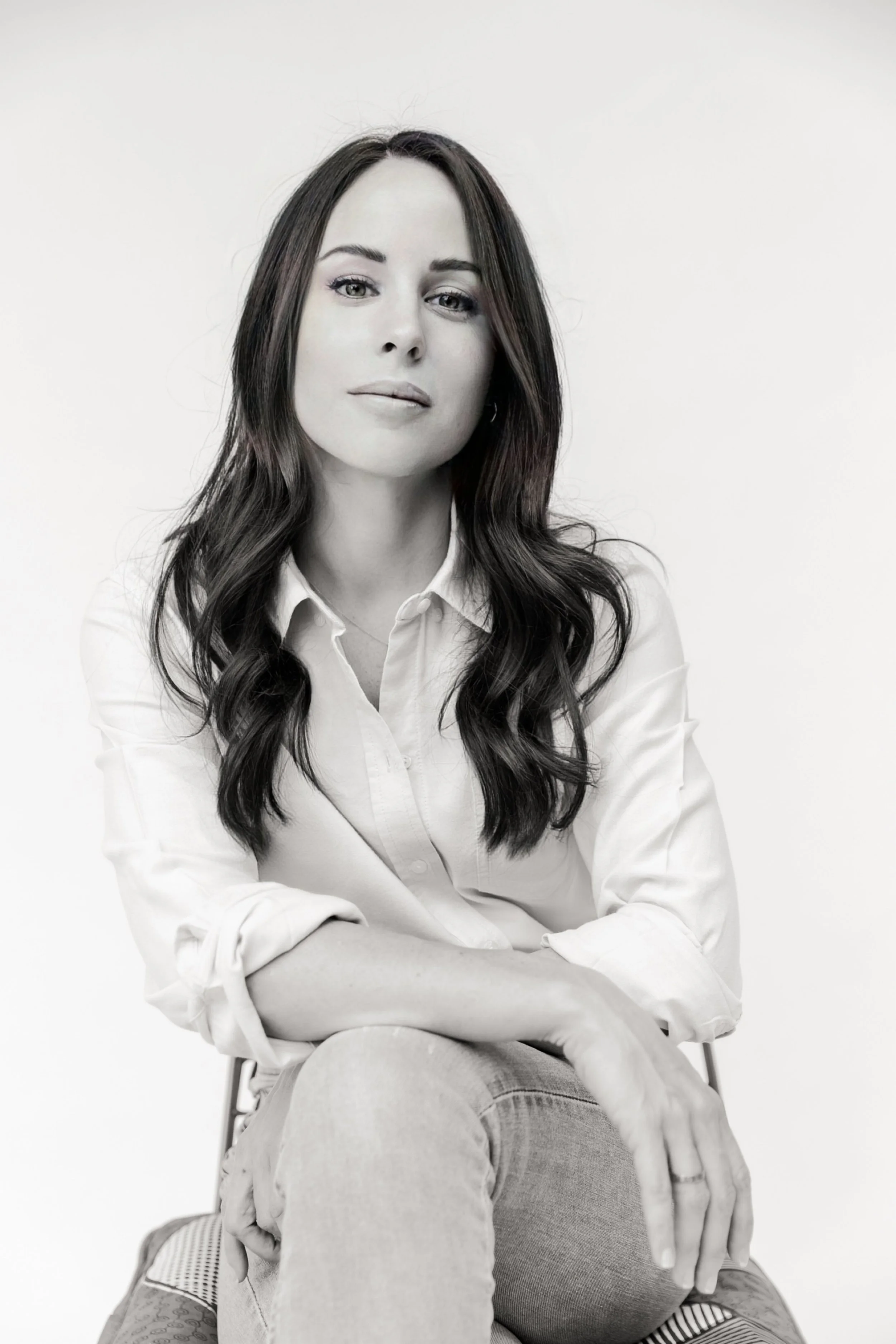 Black and white photo of a woman with long wavy hair, wearing a white button-up shirt, sitting cross-legged on a patterned cushion, with a neutral expression against a plain background.