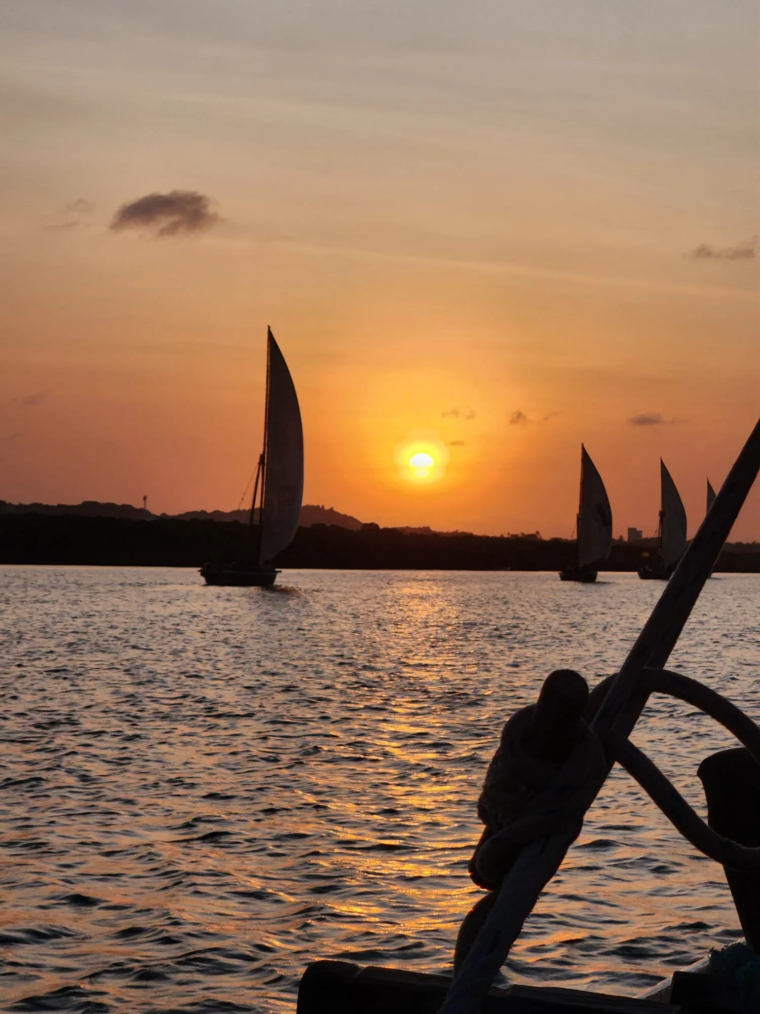 Sailboats on water during sunset with a colorful sky, a few clouds, and land in the background.
