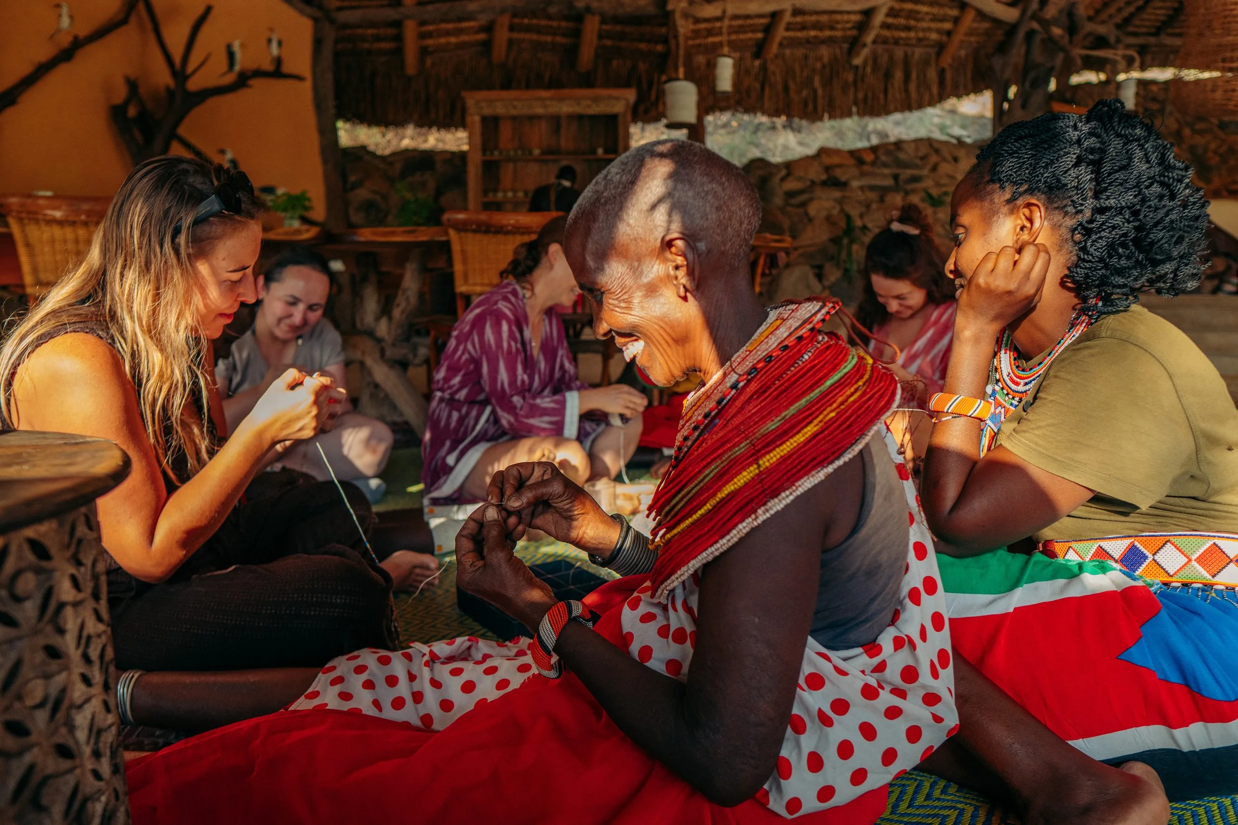 A group of women sitting and smiling inside a rustic, wooden structure. The women are of different ages and ethnicities, engaging in a shared activity, possibly sewing or crafting.
