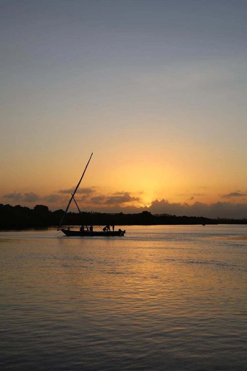 A boat with a tall sail on calm water during sunset with a silhouetted shoreline and clouds in the background.