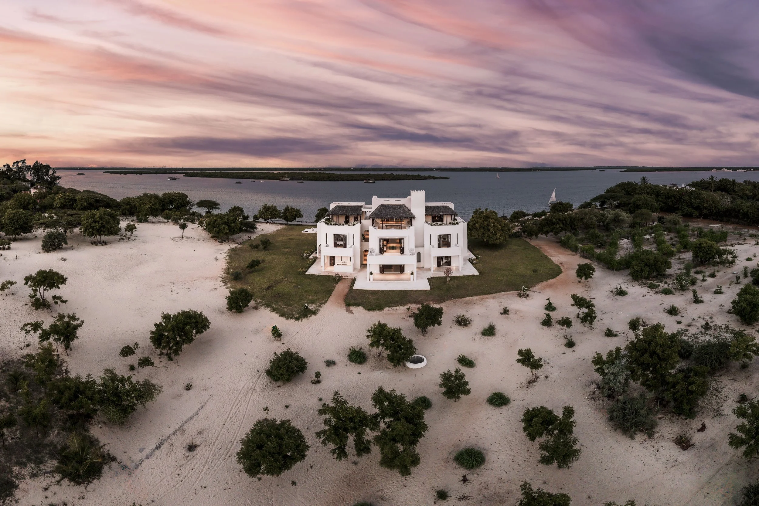 A white modern house on a sandy landscape, with trees surrounding it and a body of water in the background under a colorful sunset sky.