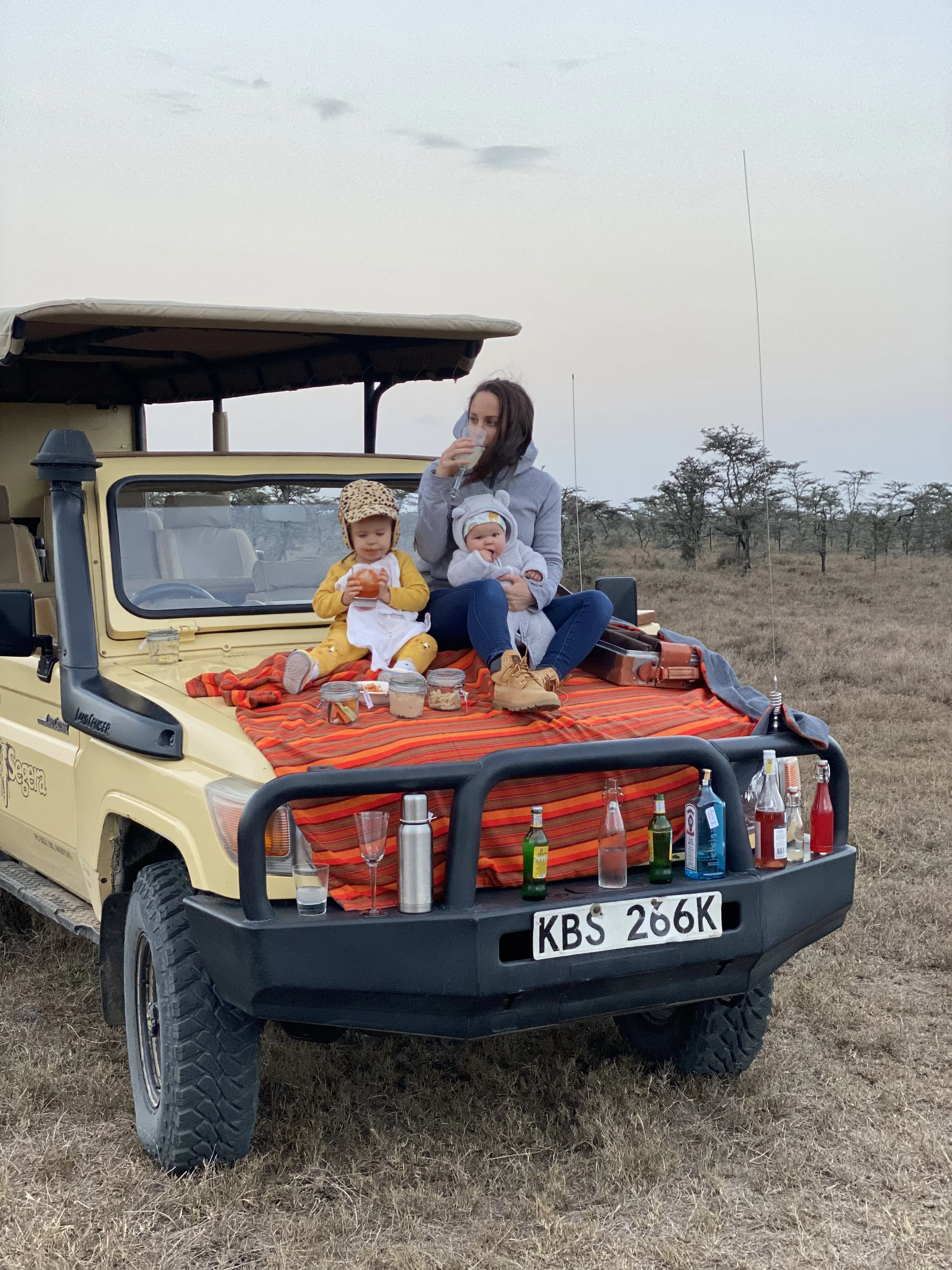 A woman and two children sitting on the hood of a yellow safari vehicle in a savannah landscape, enjoying drinks and snacks during a safari adventure at sunset.