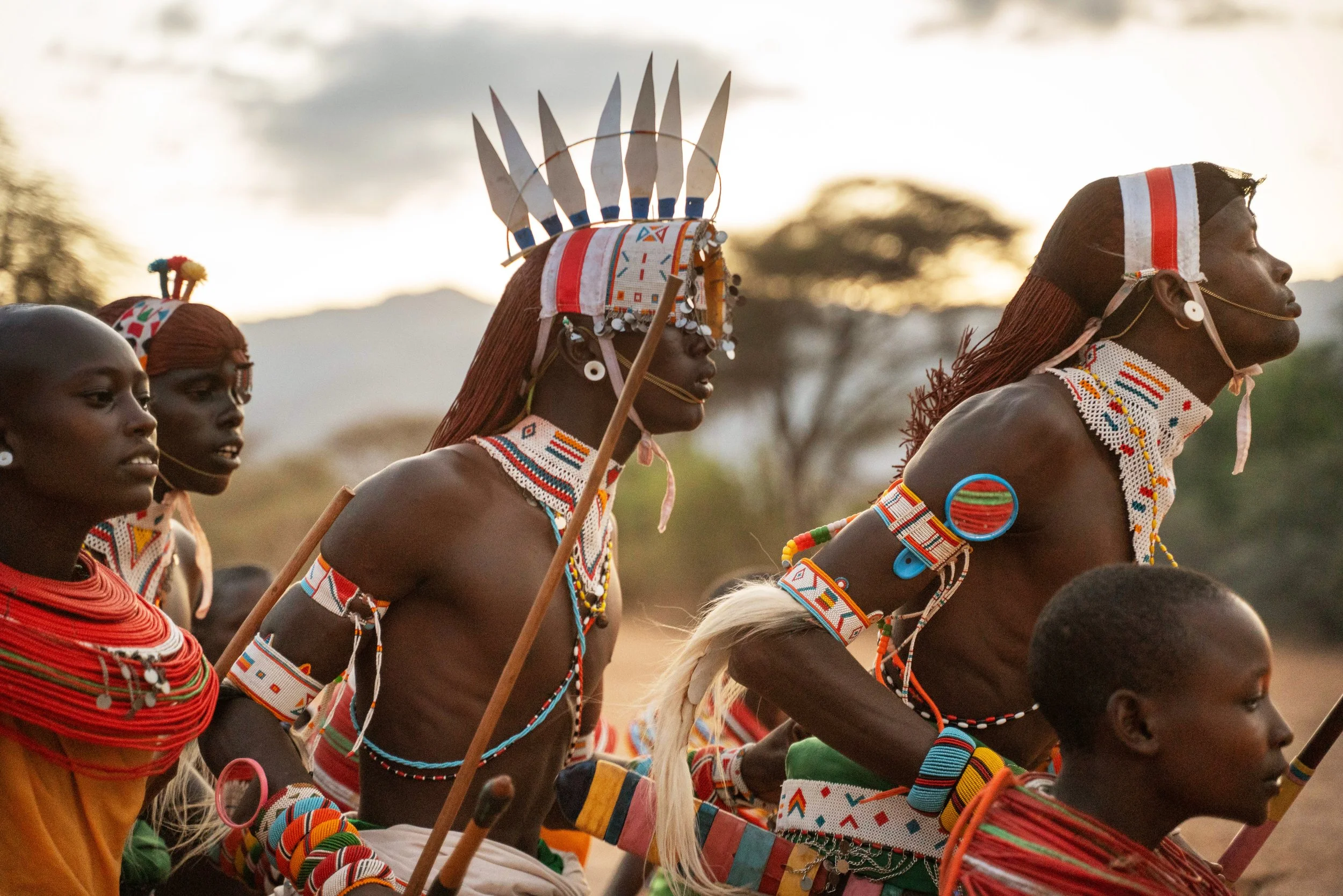 A group of Maasai people dressed in traditional attire, including beadwork, jewelry, and headpieces, participating in a cultural event outdoors at sunset.