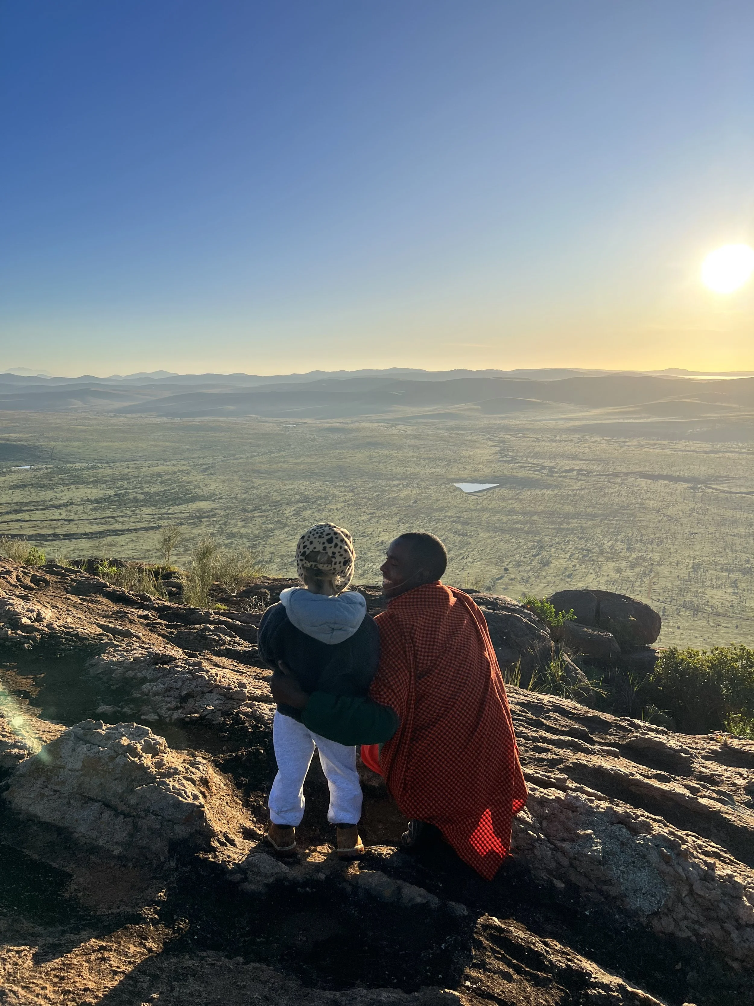 A person and a child sitting on a rocky ledge overlooking a vast landscape during sunset.