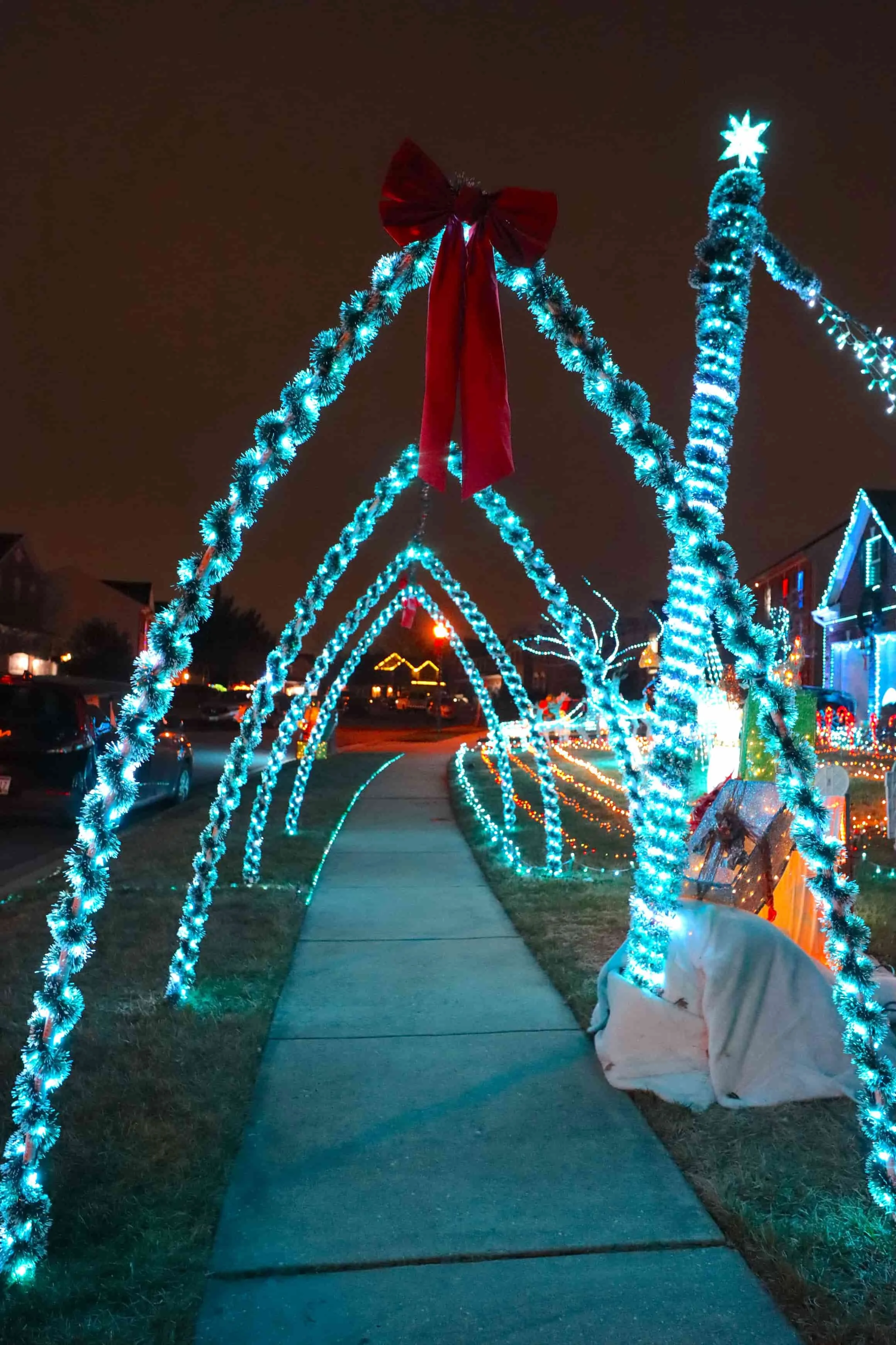 Christmas outdoor light display with illuminated arches decorated with silver tinsel, a large red bow at the top, along a sidewalk in a residential neighborhood.
