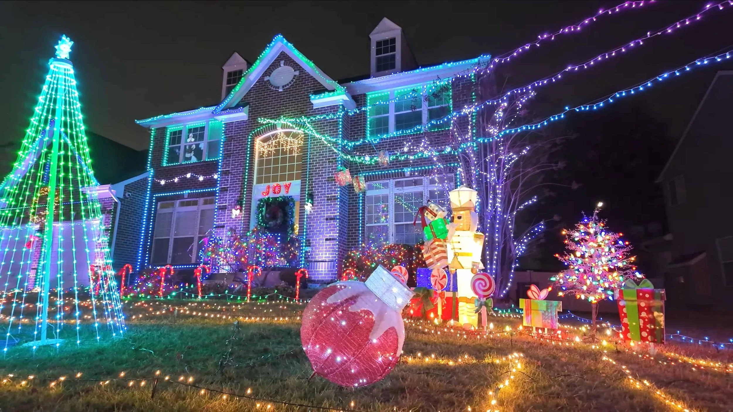 A house decorated with colorful Christmas lights, illuminated presents, a Christmas tree, light-up candy canes, and a light up nutcracker, preparing for holiday celebration.