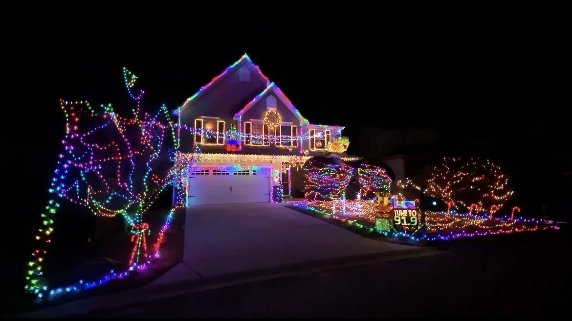 House decorated with colorful Christmas lights, including lit trees, bushes, and a wreath.