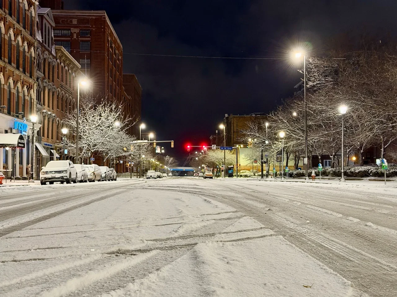 A snow-covered street in Northeast Ohio at night with parked cars, lit street lamps, and a few trees covered in snow. Buildings line the street, and traffic lights are red in the distance.