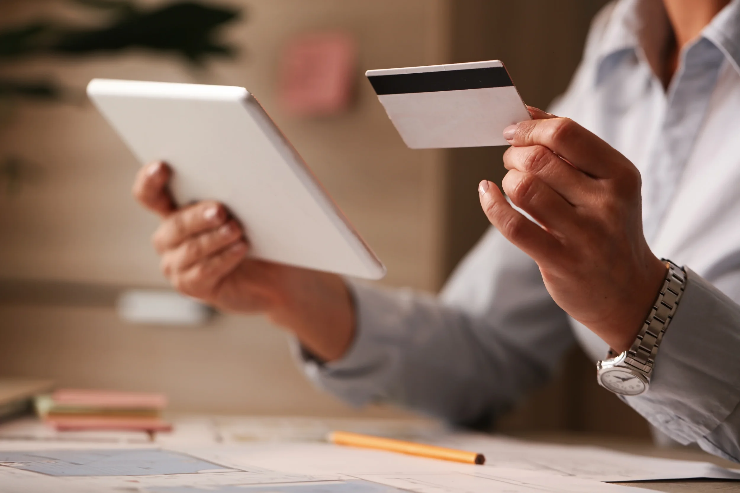 Person holding a tablet in one hand and a credit card in the other, with a desk and papers in the background.