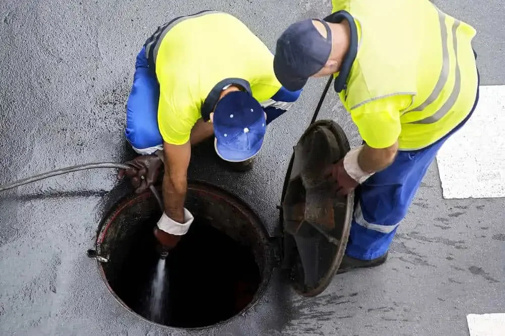 Two workers in yellow safety vests and pants, one is kneeling and the other standing, are inspecting a sewer or storm drain opening in the ground, with one pouring water or inspecting the interior.