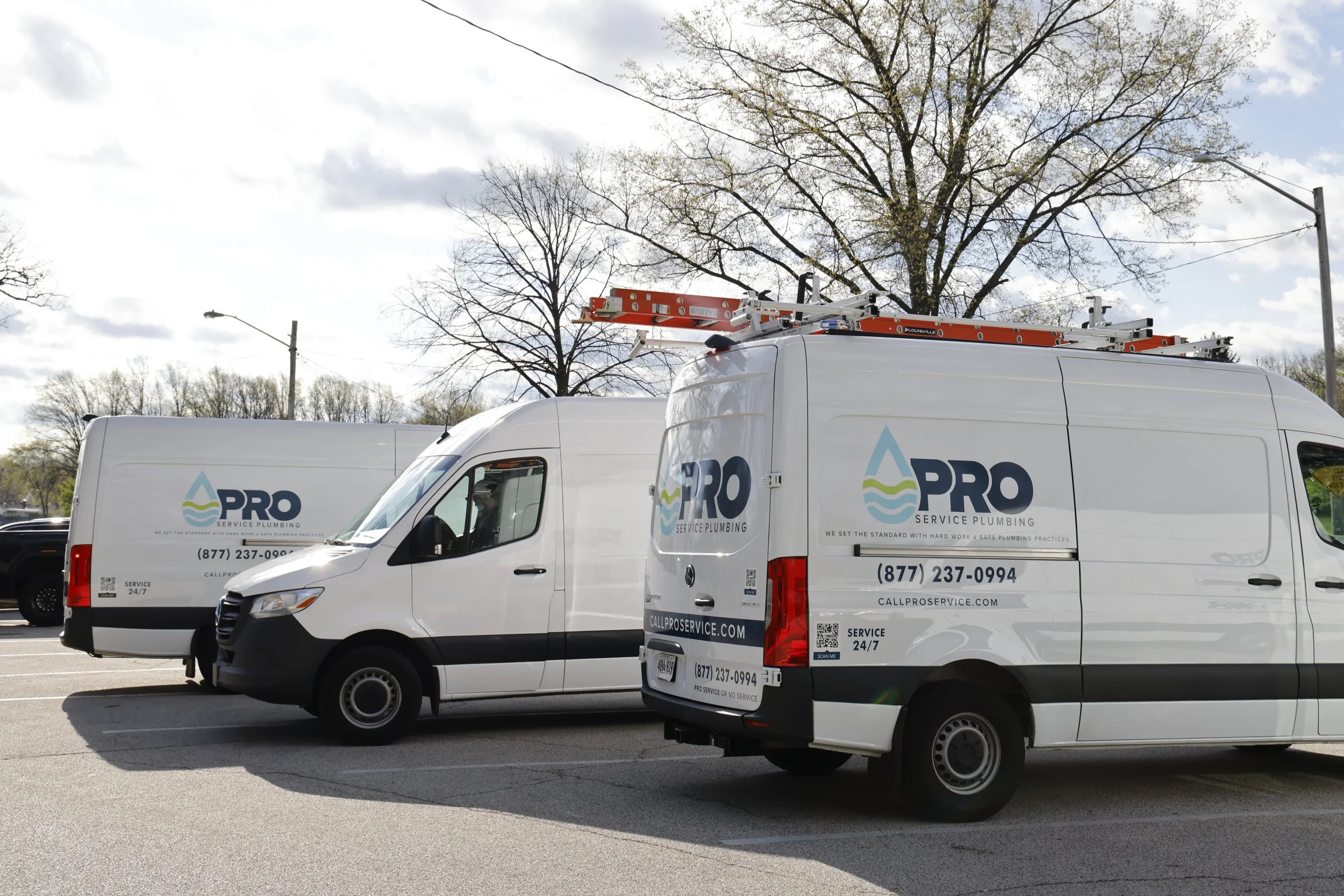 Two white service plumbing vans with the Pro Service Plumbing logo parked in a parking lot, with trees and power lines in the background.