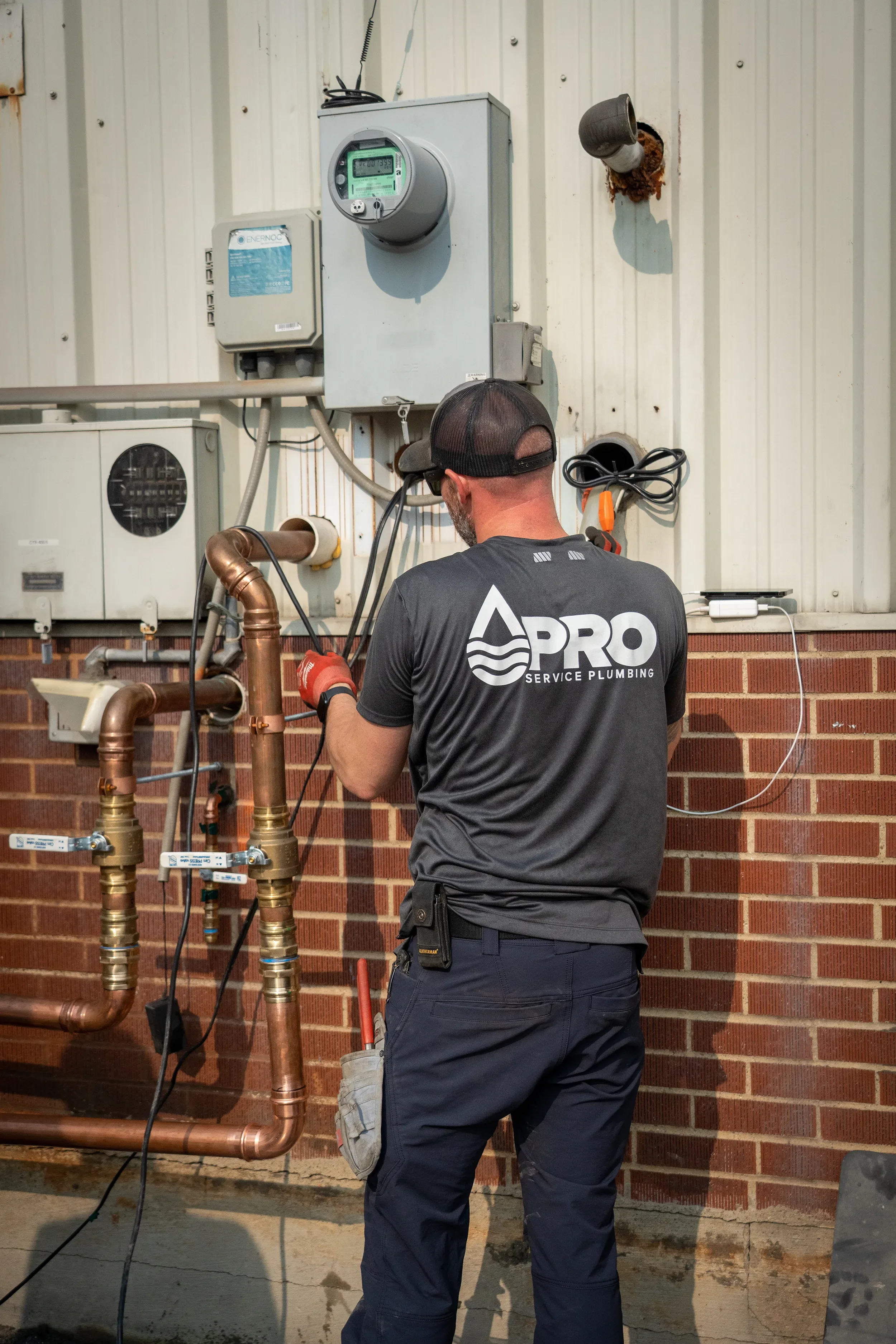 A plumber working on copper pipes outside a building, wearing a black cap and shirt with service company logo, with electrical meters and wall fixtures in the background.