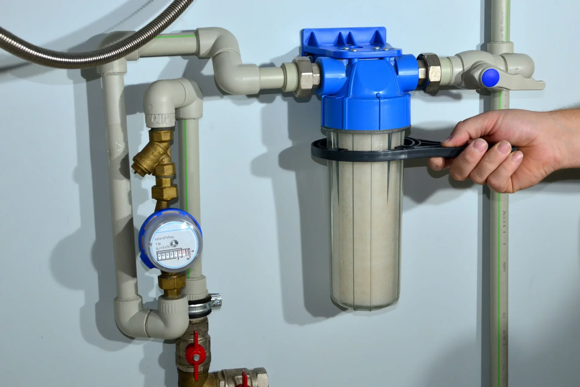 Close-up of a person tightening a large water filter with a black wrench, surrounded by plumbing pipes and a water meter.