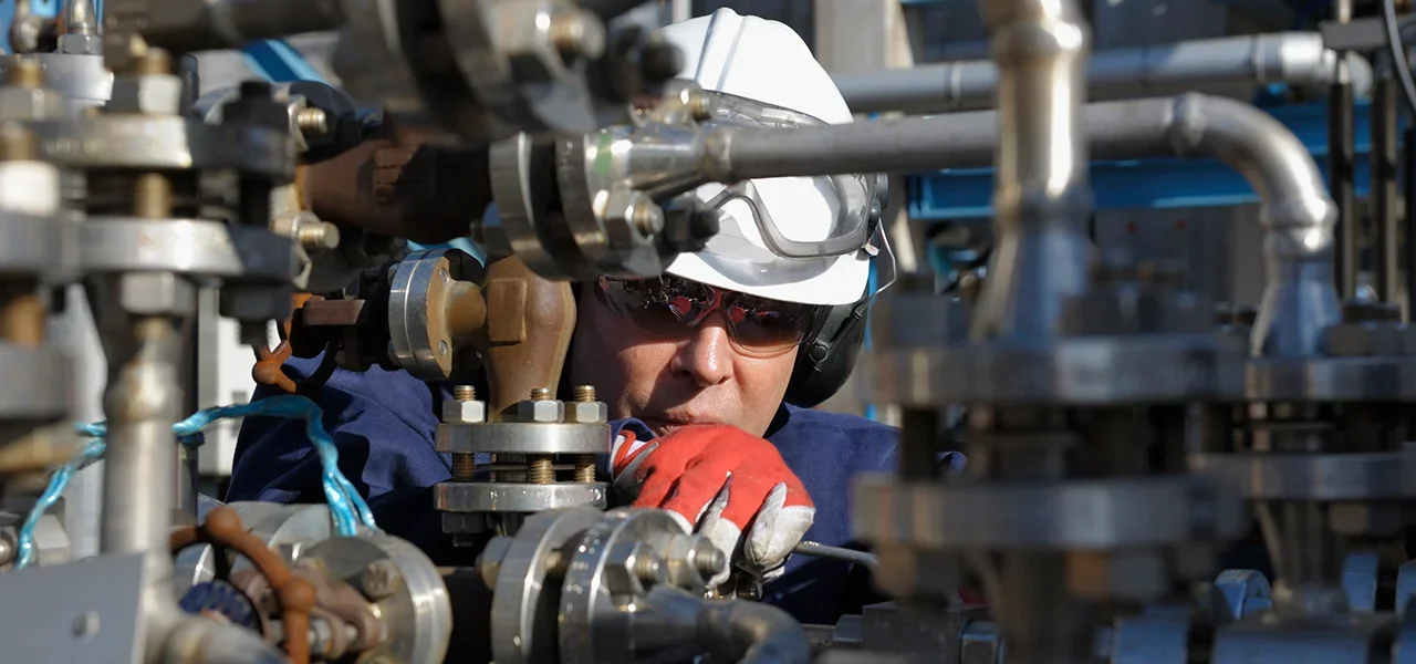 A worker wearing a white safety helmet, goggles, and orange gloves working on industrial pipes and valves.