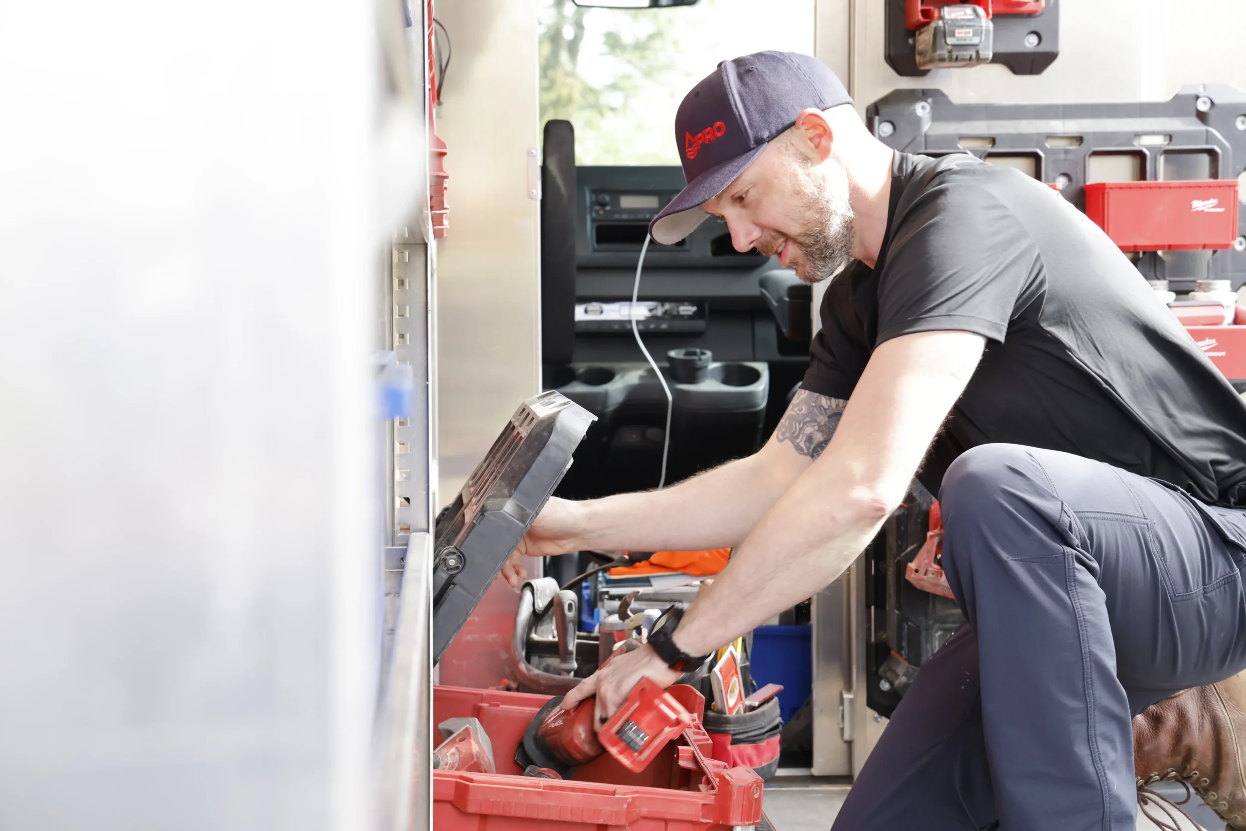A man in work attire kneeling beside an open red tool box while organizing tools inside it.