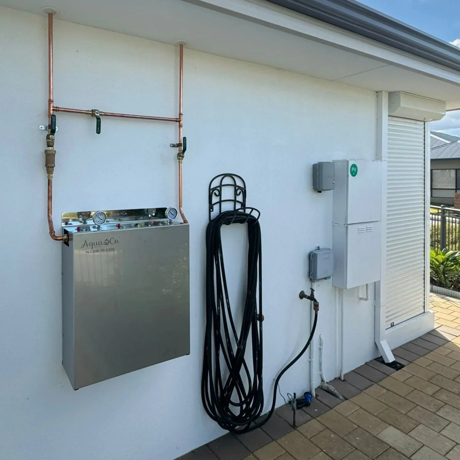 Outdoor wall with plumbing, electrical boxes, water hose, and a black metal stand, adjacent to a shaded patio and a residential neighborhood.