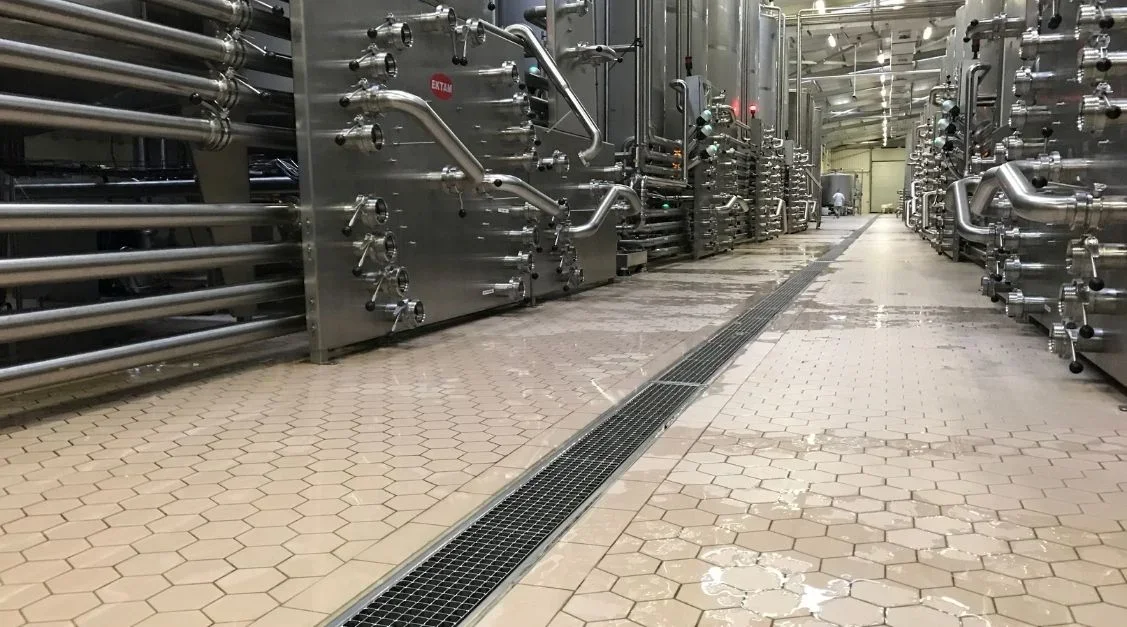 Industrial facility with large stainless steel pipes and tanks, tiled floor, and a drainage grate in the center.