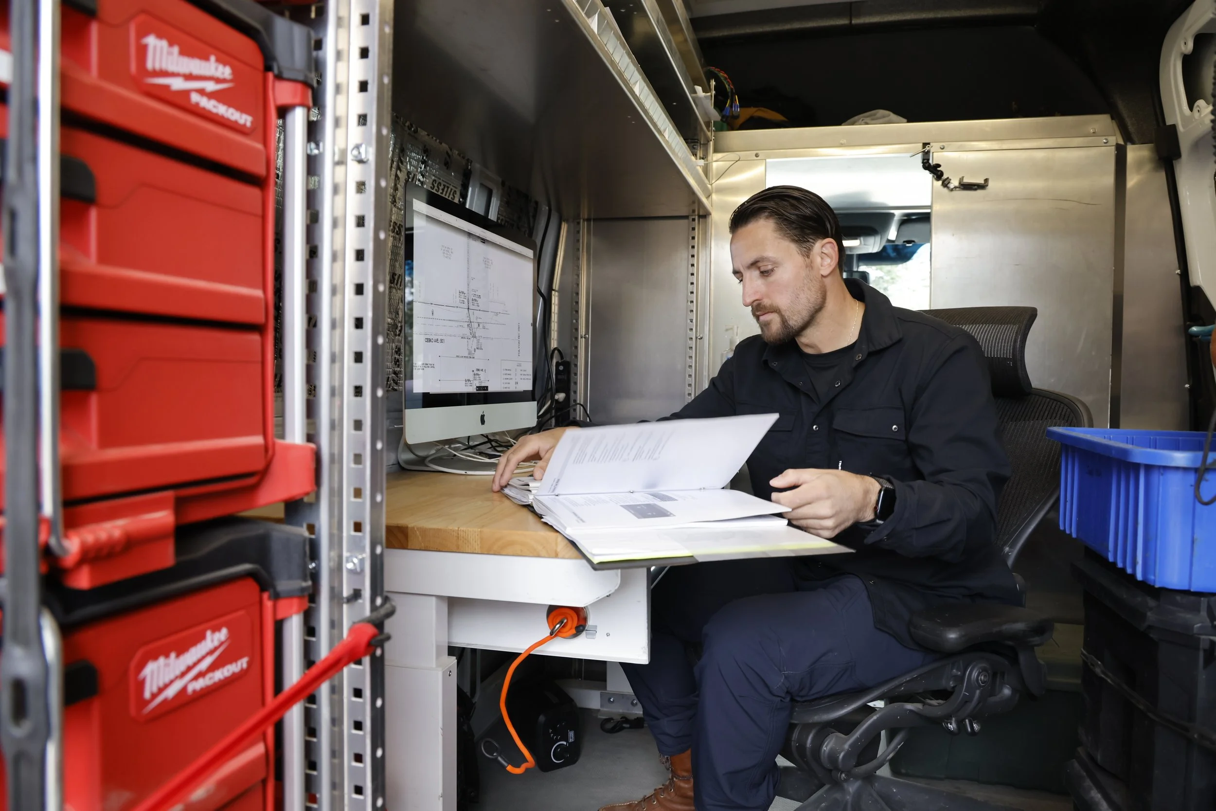 A man working on a computer inside a mobile workspace, with tools and storage bins around him.