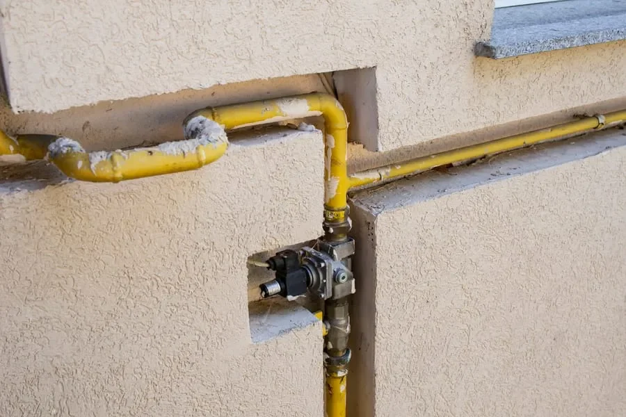 Yellow gas pipe with a valve and mechanical component mounted on a beige stucco wall of a building.