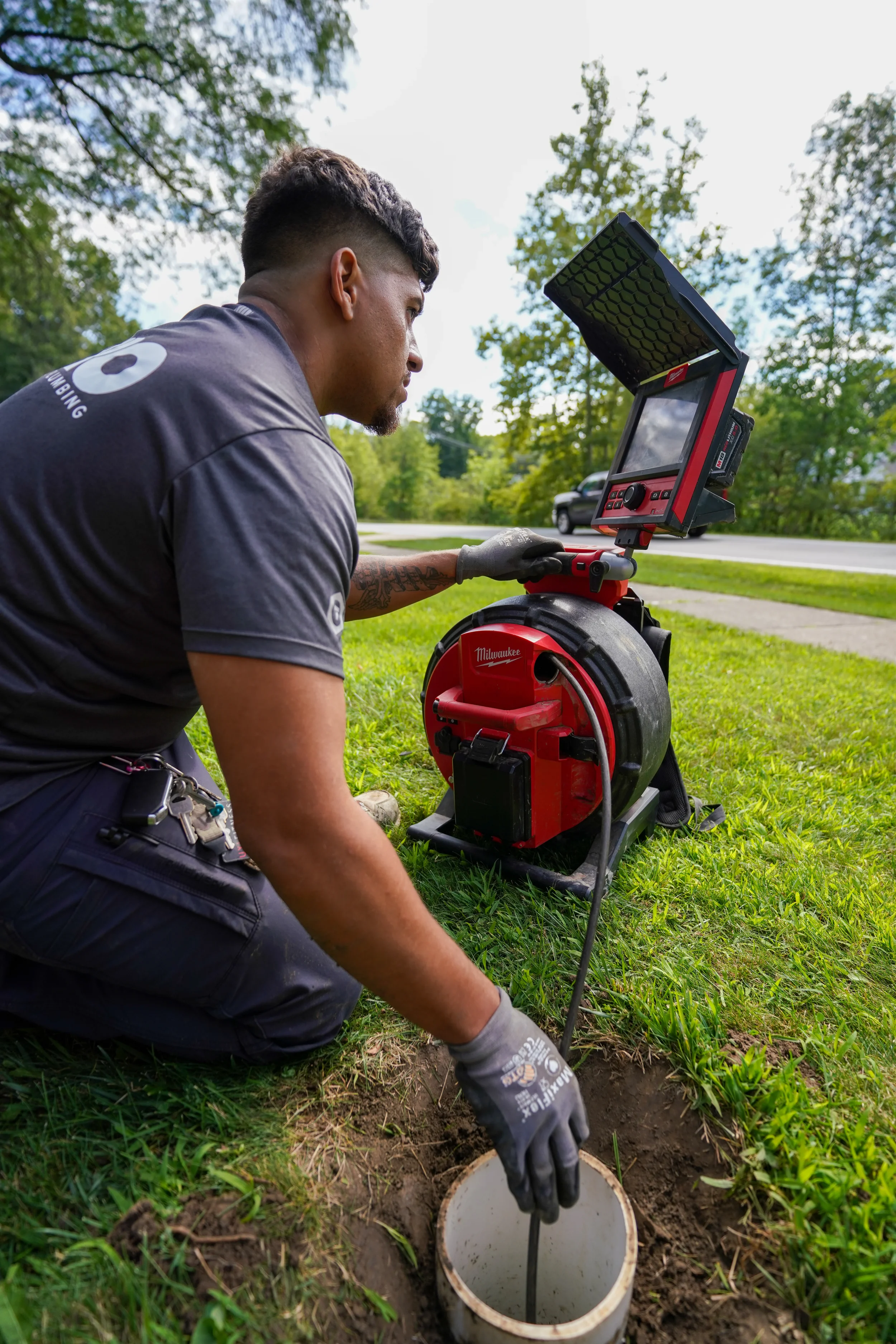 A utility worker kneeling on grass, installing a pipe into the ground with the help of a sewer camera and inspection equipment, outdoors near a neighborhood street.