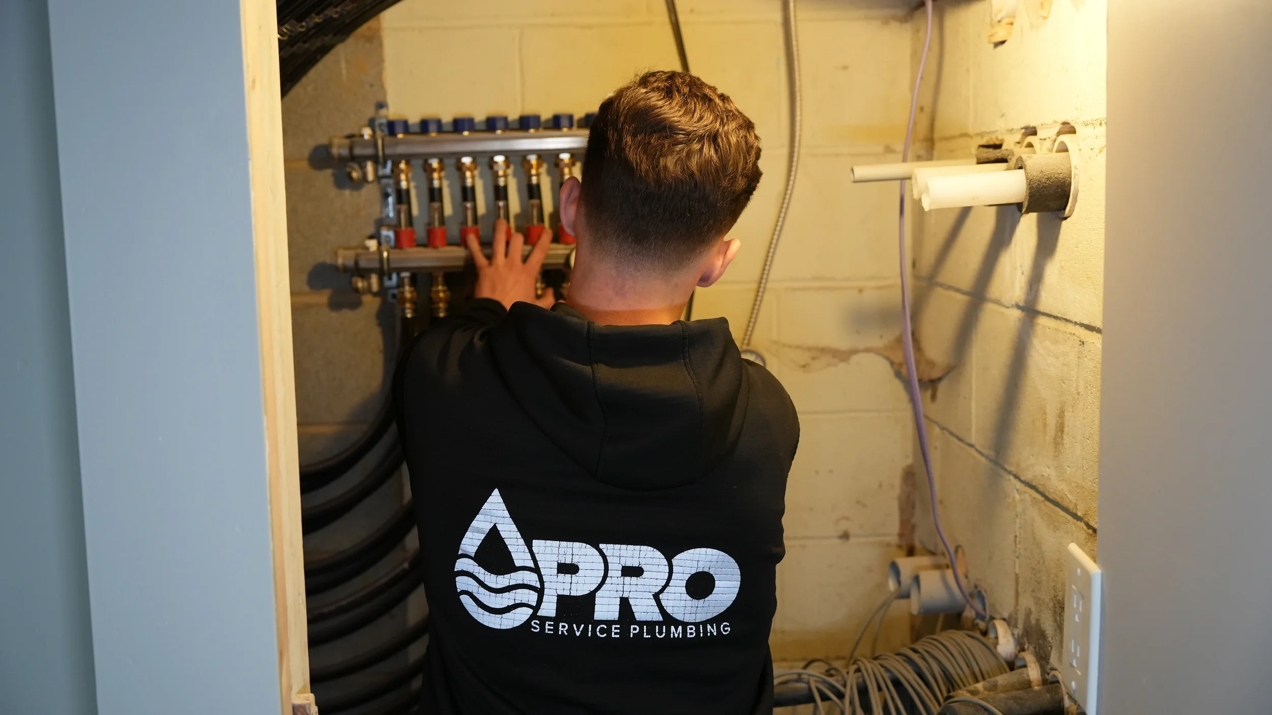 A plumber working on a plumbing manifold inside a utility closet, wearing a black hoodie with 'PRO Service Plumbing' logo on the back.