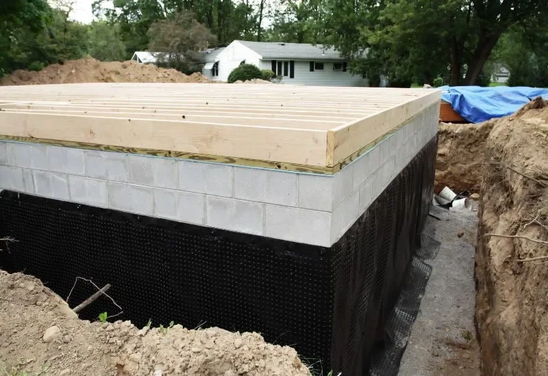 Foundation of a building under construction, with layers of concrete blocks, a black waterproof barrier, and a wooden frame on top, surrounded by dirt and construction site materials.