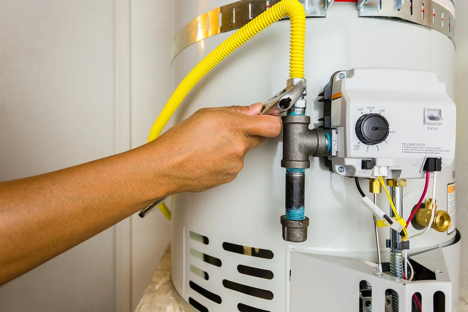 A person repairing or maintaining a white tankless water heater by tightening a connection with a wrench. The water heater has yellow and black hoses, a control panel with a dial and an indicator light, and various wires and components.