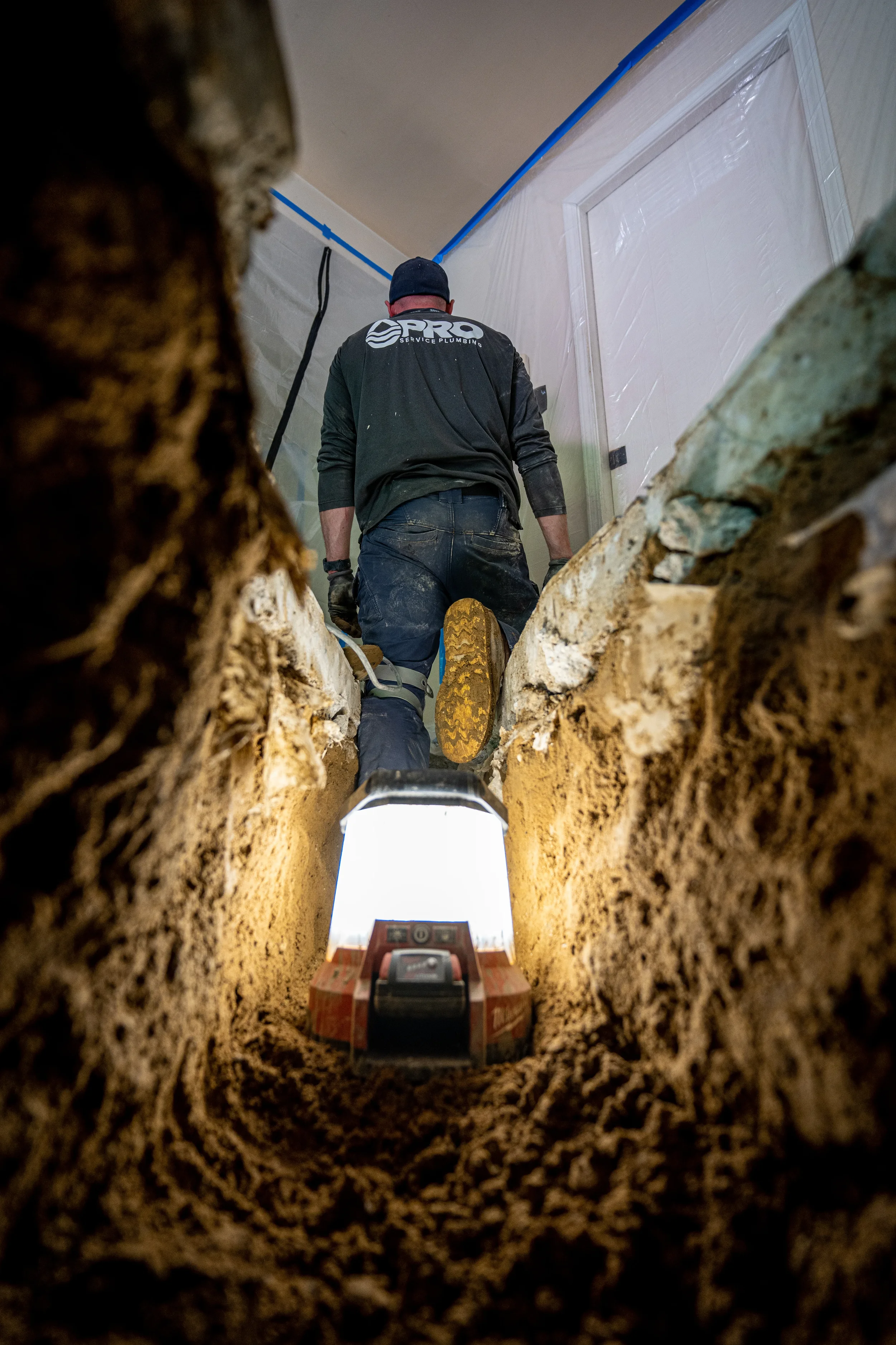A Pro Service plumber in black clothing in a narrow trench, working on a wall near a door. The photo is taken from inside the trench, showing the worker from below with a level placed in the trench for leveling.