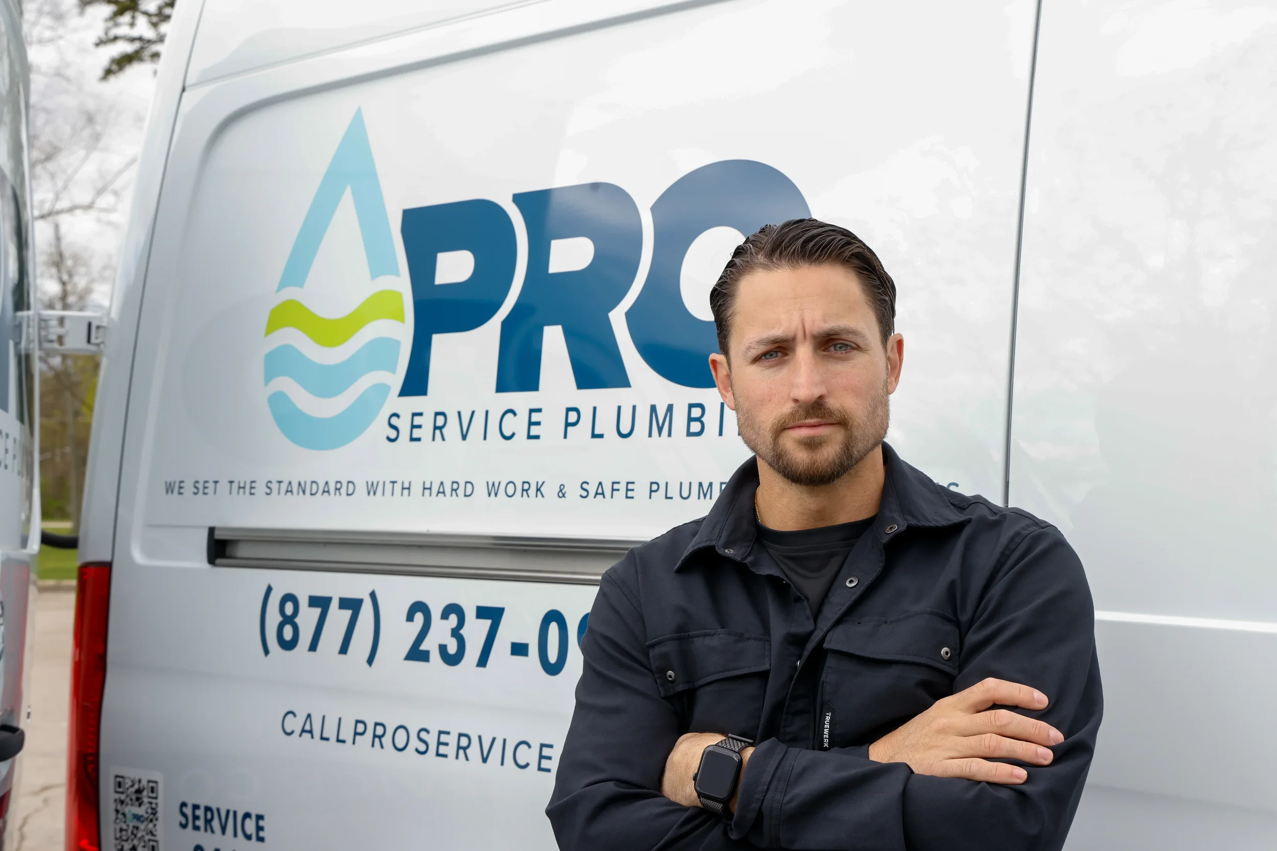 A man with a beard and short hair standing with arms crossed in front of a Pro Service Plumbing van.