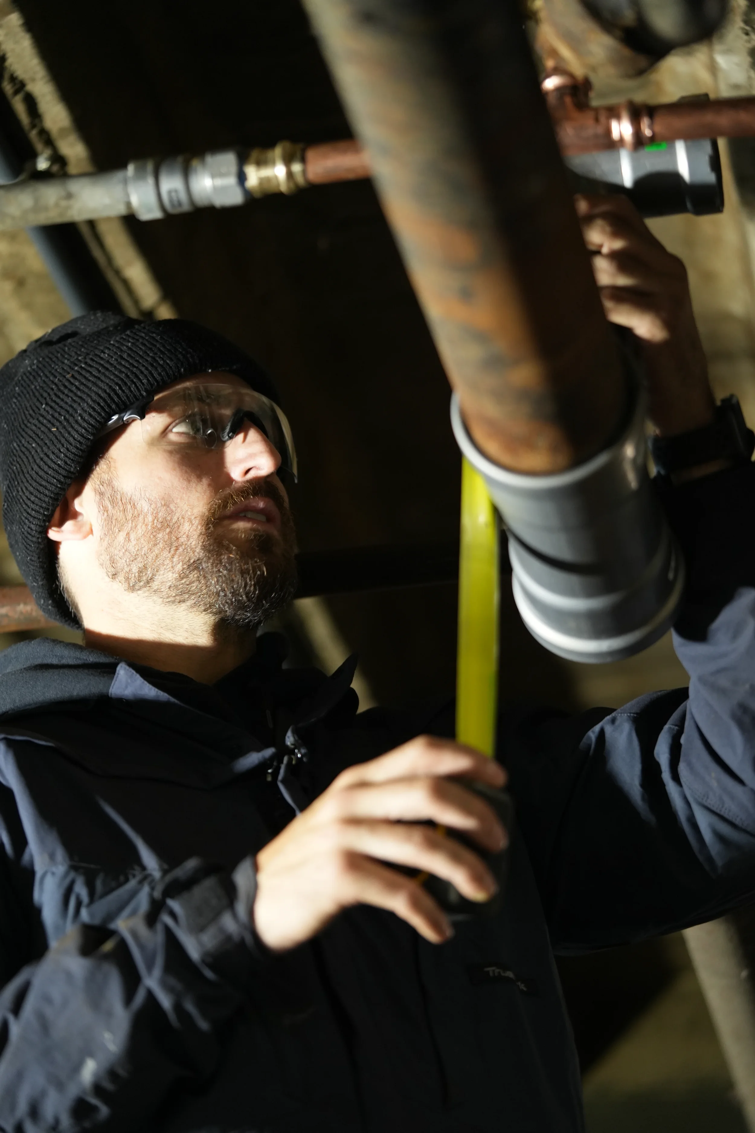 A man in safety glasses, a black beanie, and a dark jacket using a measuring tape to measure a pipe in an underground setting.