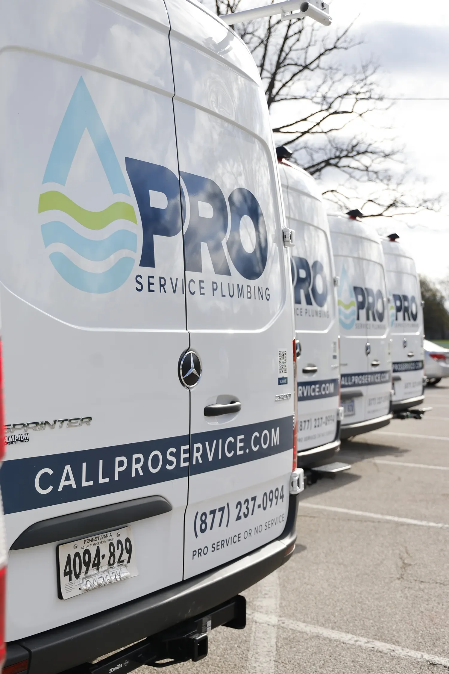 A row of white service vans with PRO Service Plumbing logos parked on a lot, with a tree and cloudy sky in the background.