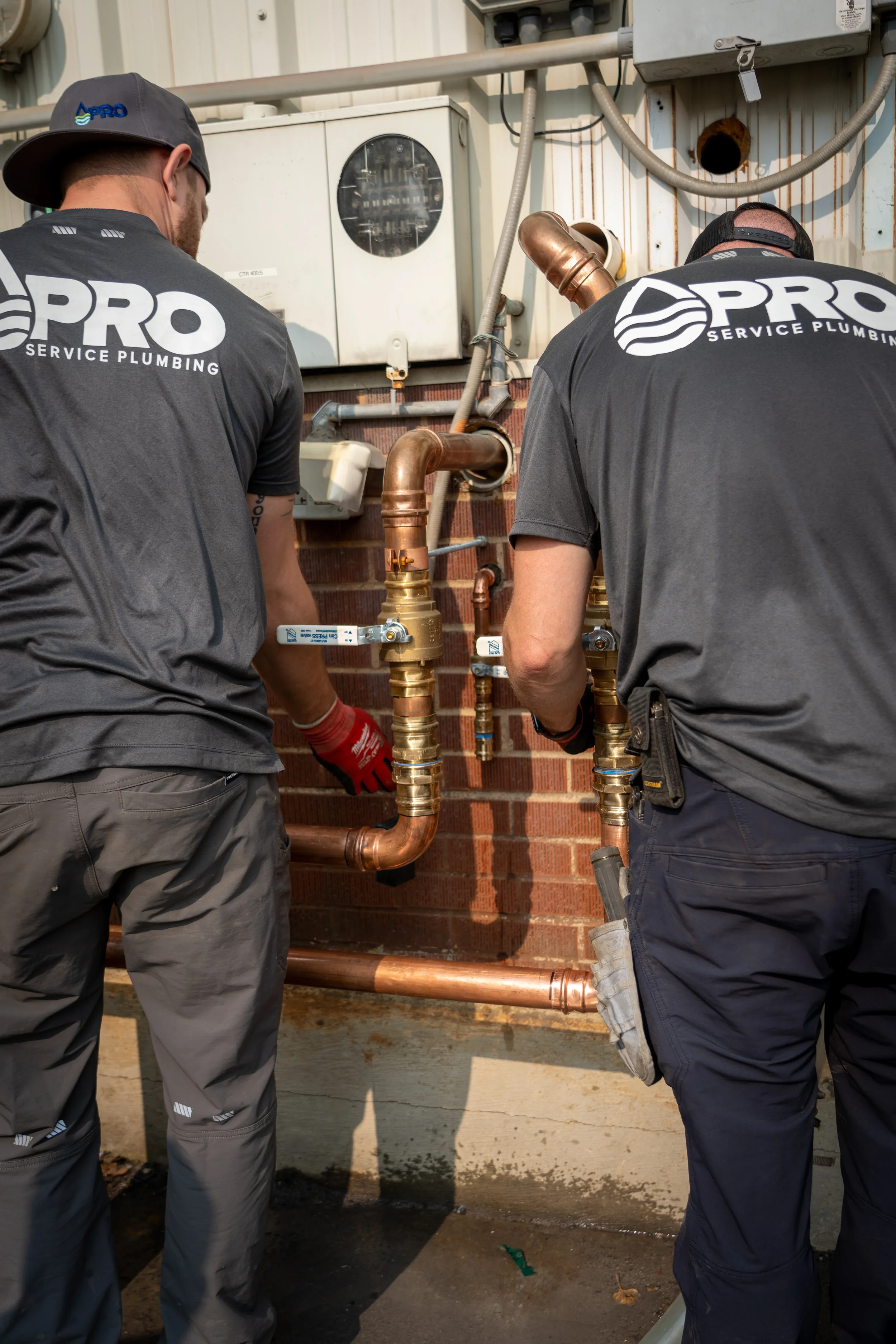 Two plumbers working on copper pipes and fittings outside a building, with electrical panels and conduits in the background.