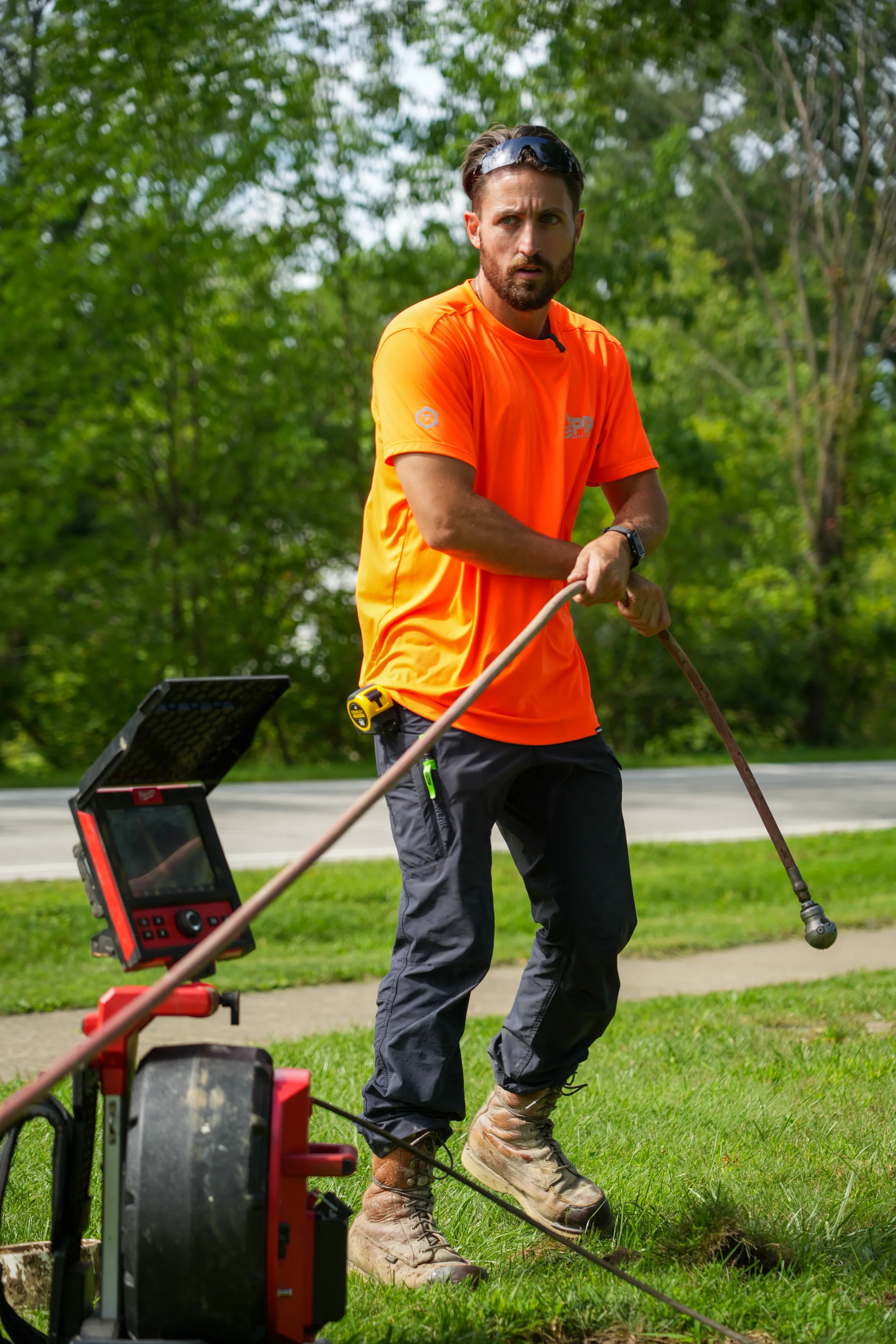 A man wearing an orange shirt, gray pants, and sturdy boots is operating a ground-penetrating radar device on a grassy area with trees in the background.