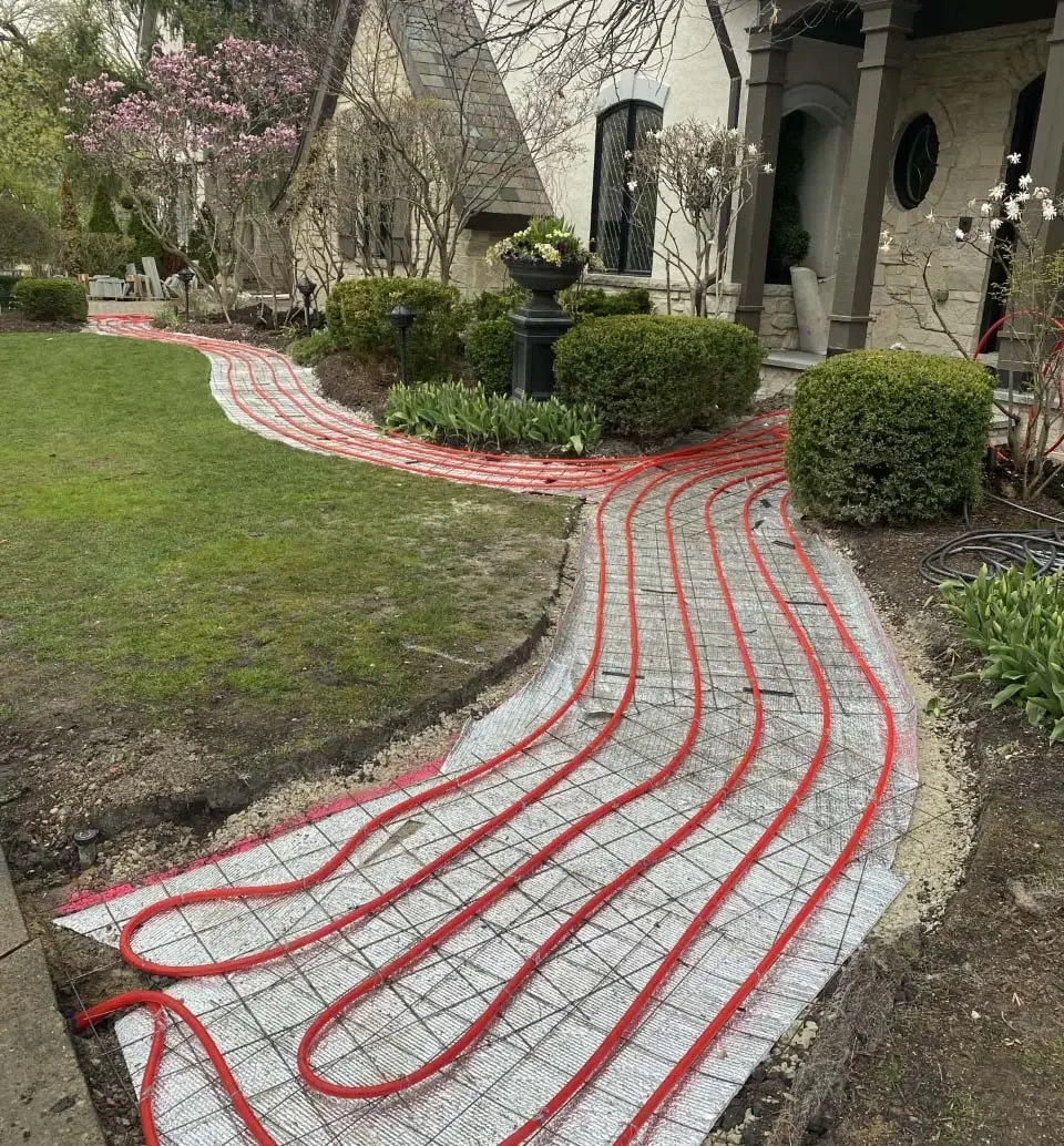 A walkway under construction with embedded red tubing for radiant floor heating, surrounded by grass, bushes, and flower beds near a house.