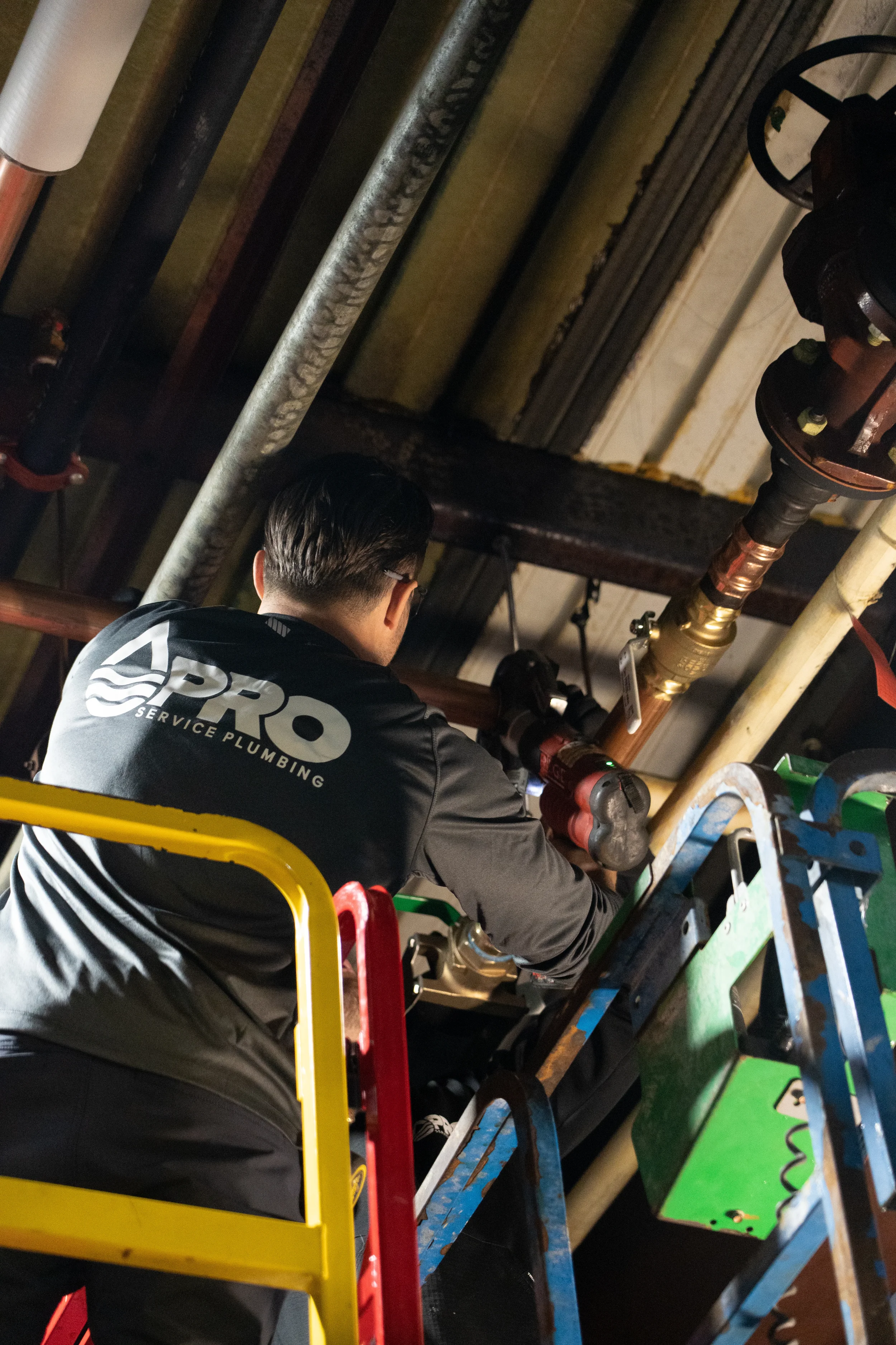 A plumber working on pipes in a building ceiling, wearing a black jacket with the words 'Pro Service Plumbing' on the back, on a yellow scissor lift.