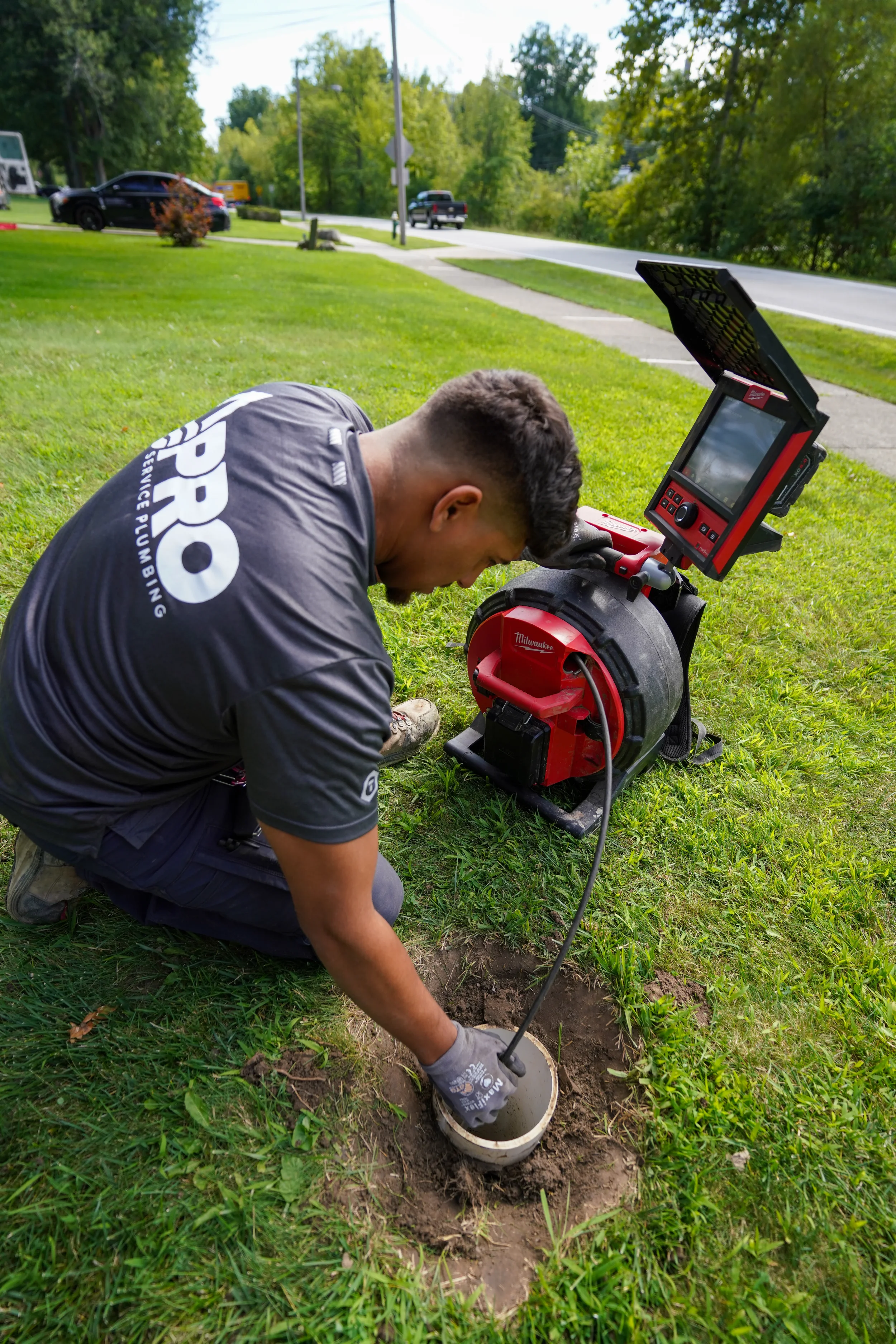 A man kneeling on grass and performing a plumbing or sewer inspection using a sewer camera next to a red Milwaukee drain camera device.